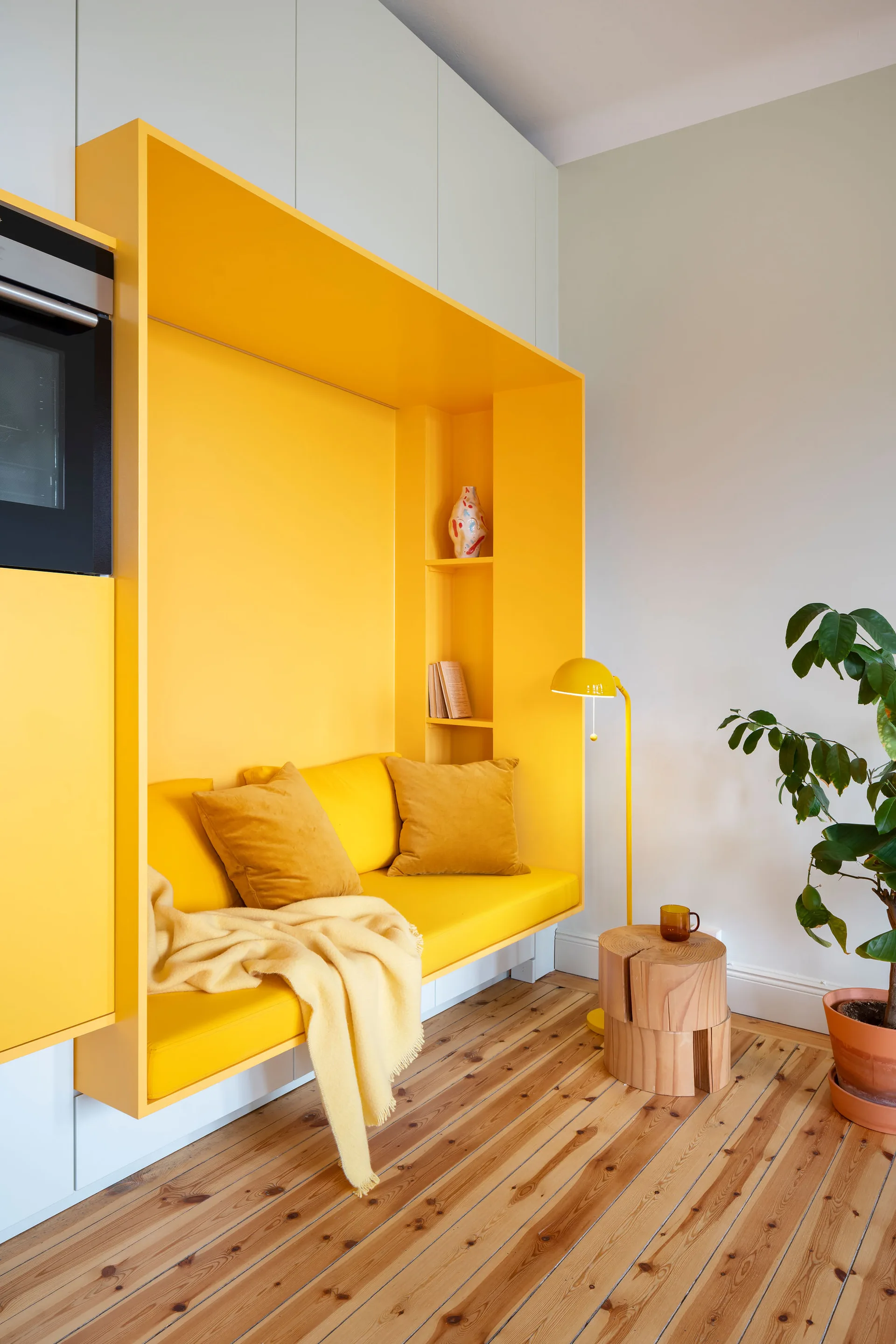 A bright yellow built-in reading alcove with pillows is set into white wall cabinets next to a wood log side table and a potted plant.