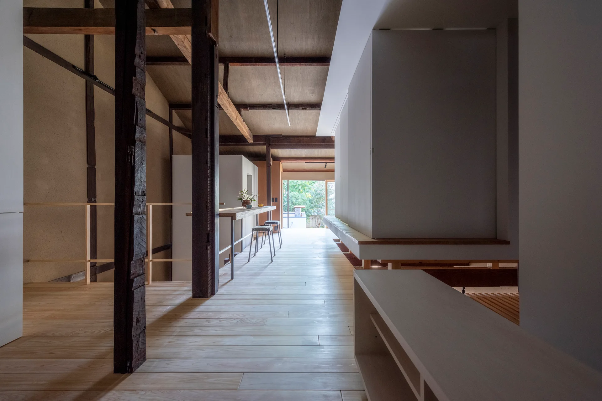 A minimalist corridor featuring light wood plank flooring, dark timber support pillars, exposed ceiling beams, and a narrow wooden bar.
