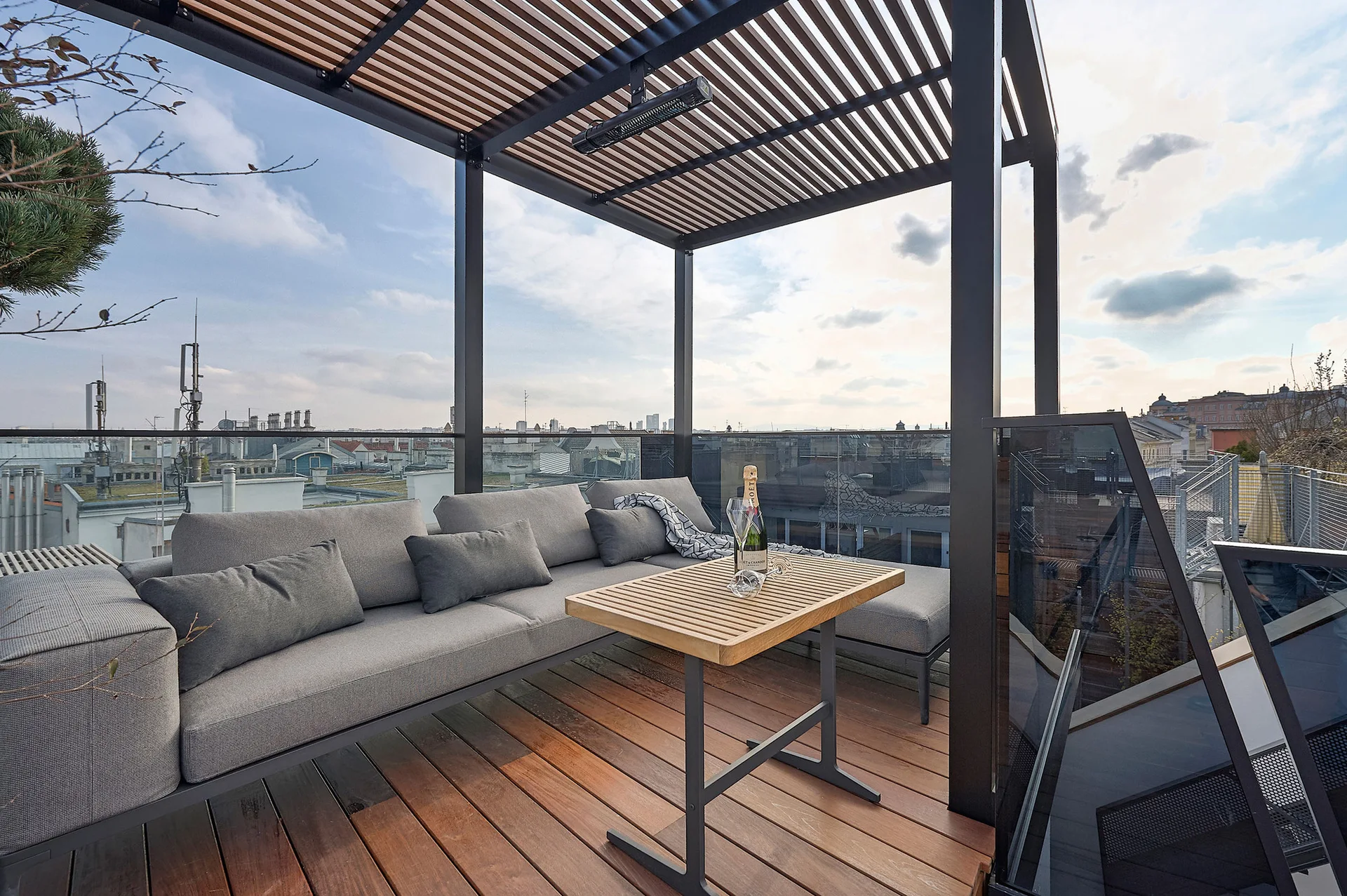 A rooftop terrace features a gray sectional sofa, a wooden coffee table, and a slatted pergola overhead, overlooking a city skyline.