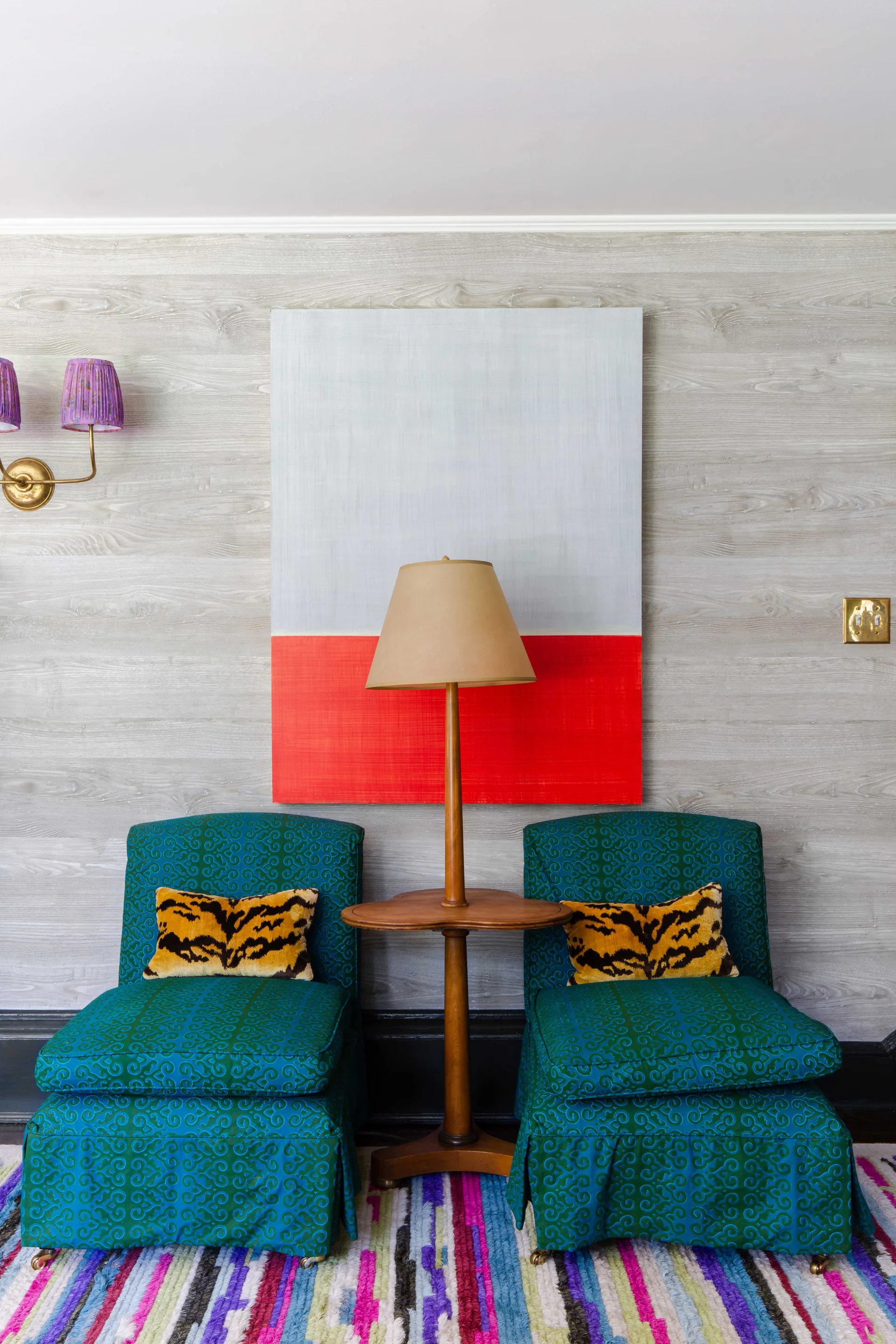 Two teal patterned armchairs sit symmetrically beside a wooden table on a colorful striped rug under a large red and grey painting.