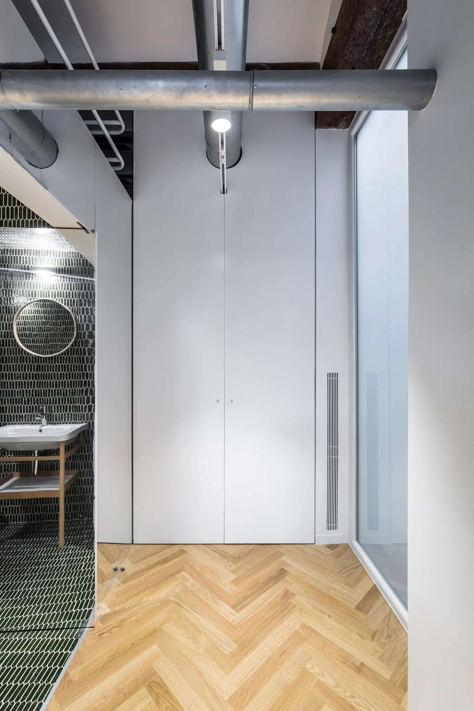 A minimalist hallway features light oak herringbone floors, white tall built-in closets, exposed ceiling ductwork, and an adjacent dark tiled bathroom.