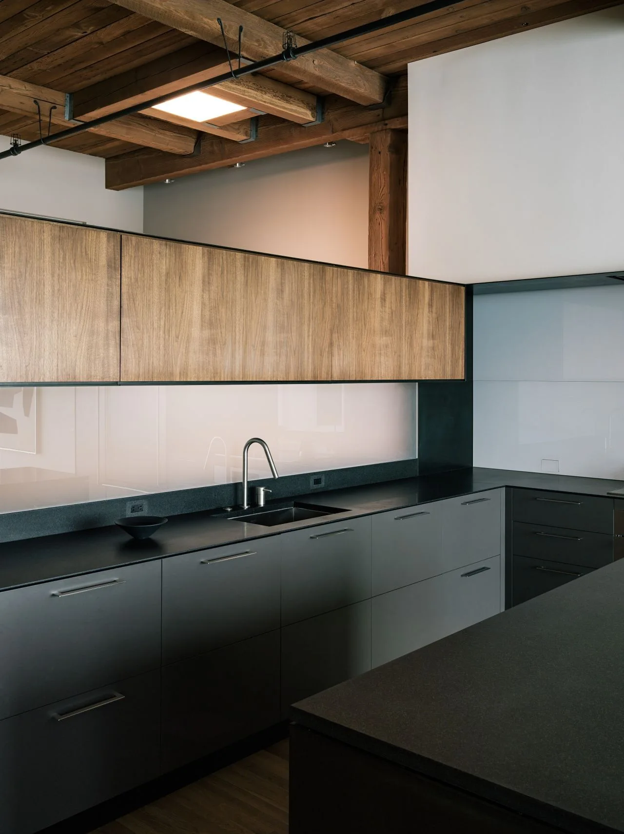 A modern loft kitchen features dark matte black lower cabinets, integrated sink, flat-panel wood upper cabinets, and exposed ceiling beams.