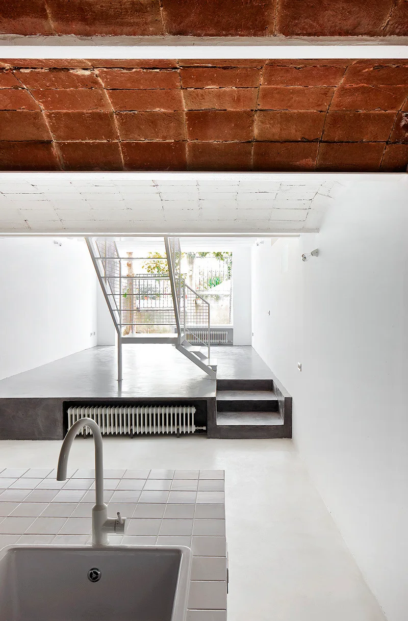 A split-level room features a tiled kitchen counter in the foreground, a white metal staircase, and an exposed brick vaulted ceiling.
