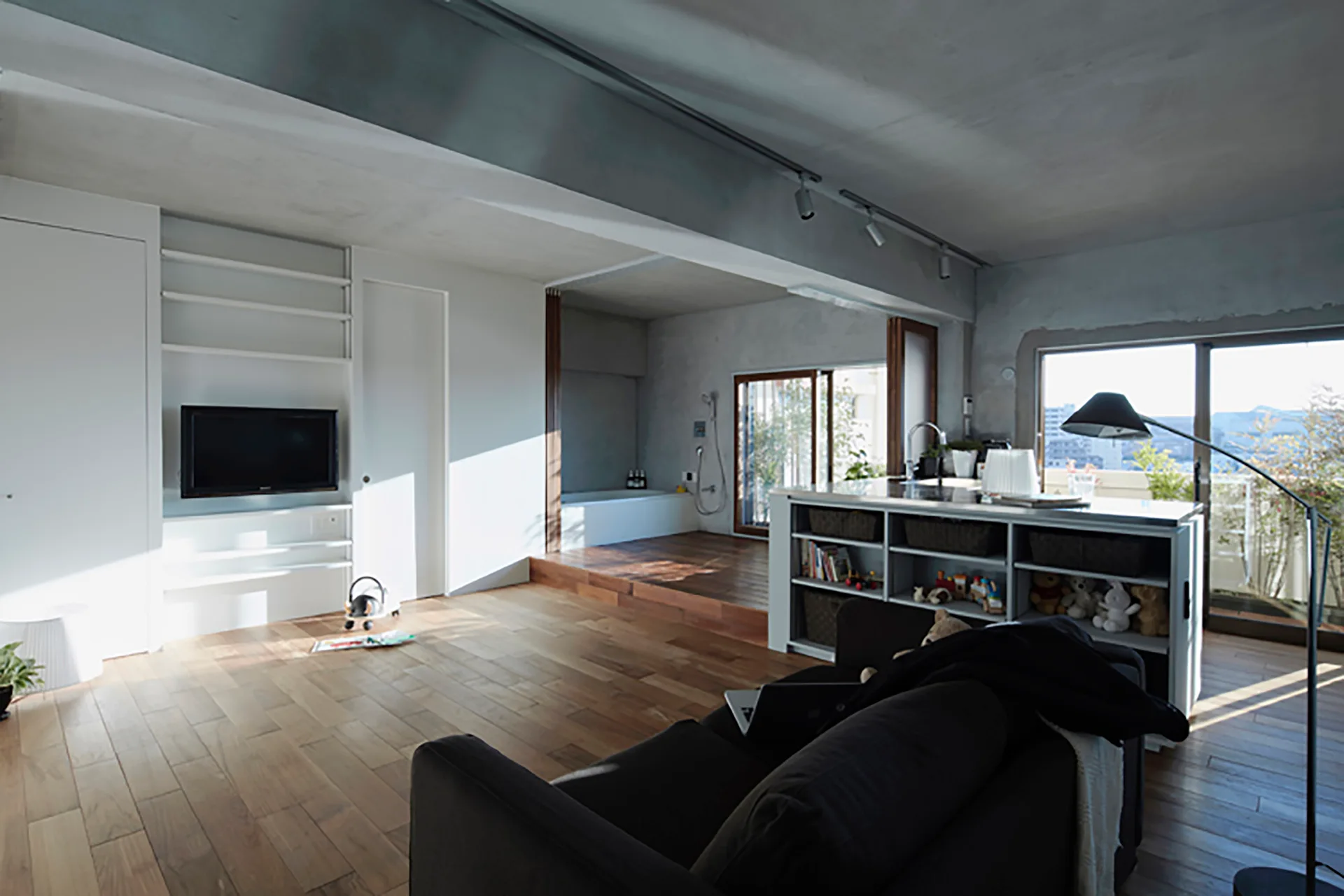 An open-concept living room with a dark sofa, a kitchen island with built-in shelves, a wooden floor, and an exposed concrete ceiling.