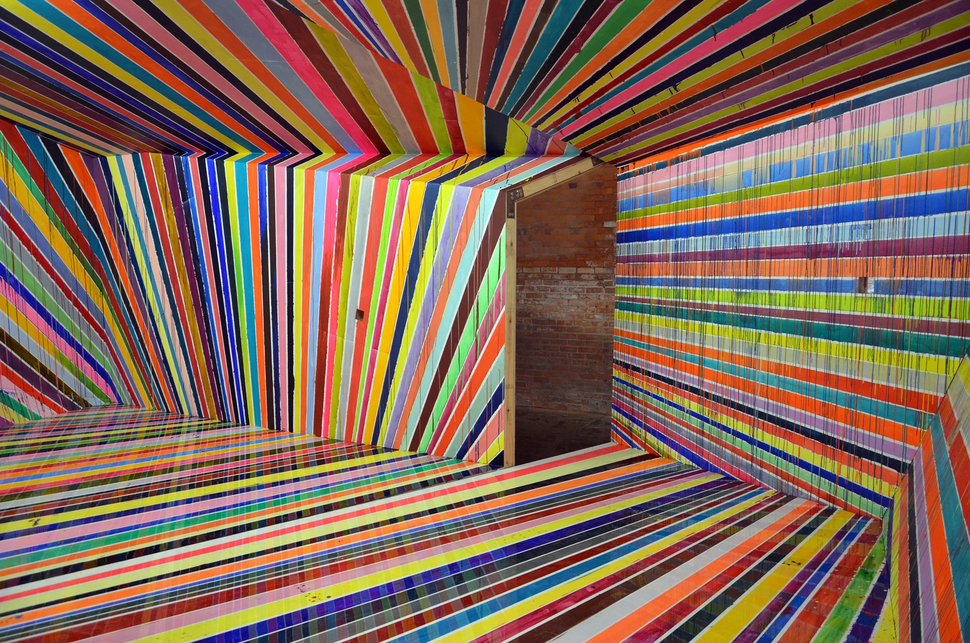 An interior space painted entirely with vibrant multicolored stripes on the walls, floor, and ceiling, featuring a doorway that opens to exposed brick.