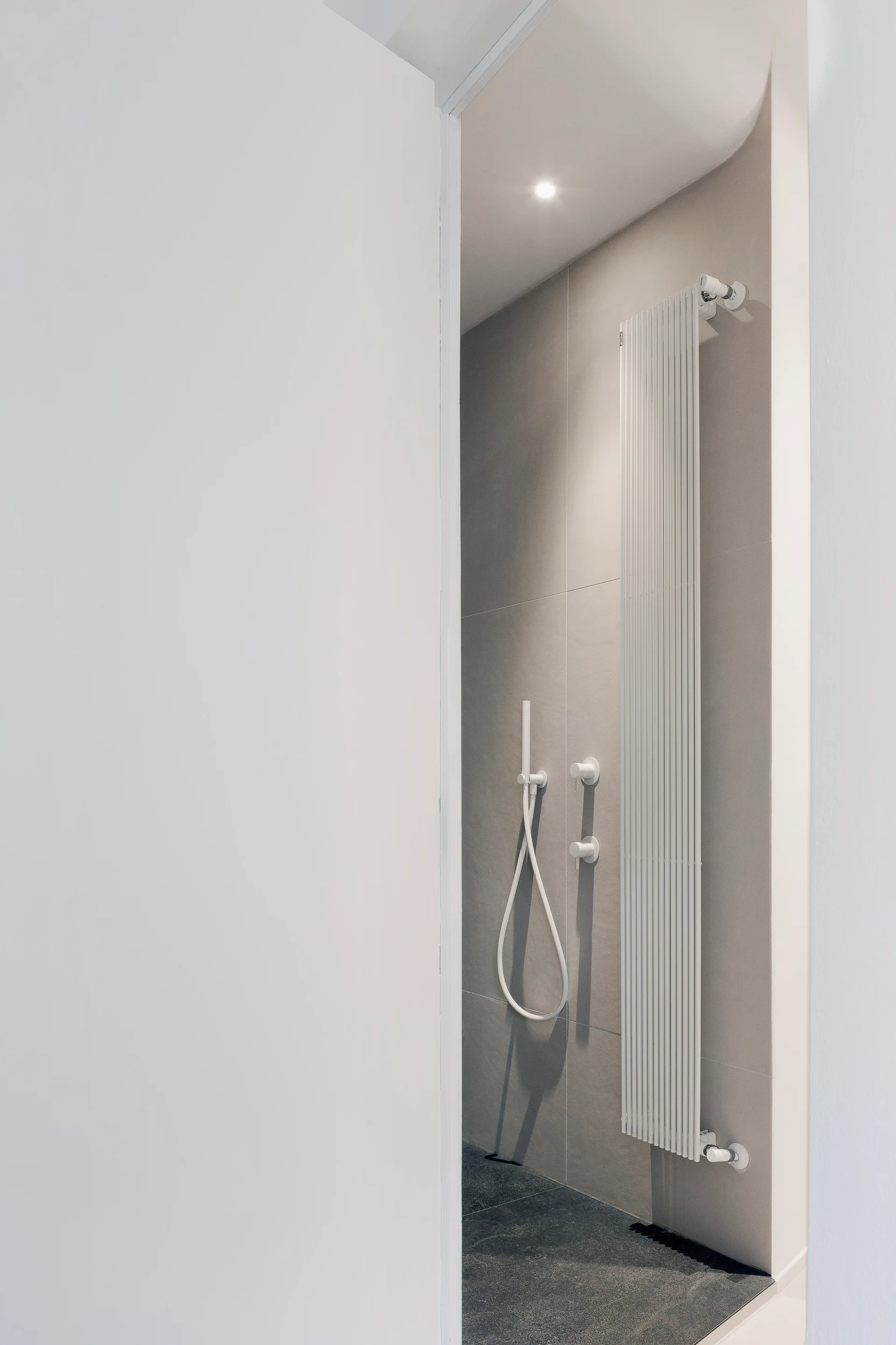 A view through a white doorway into a minimalist bathroom featuring grey wall tiles, a white vertical radiator, and a white shower fixture.