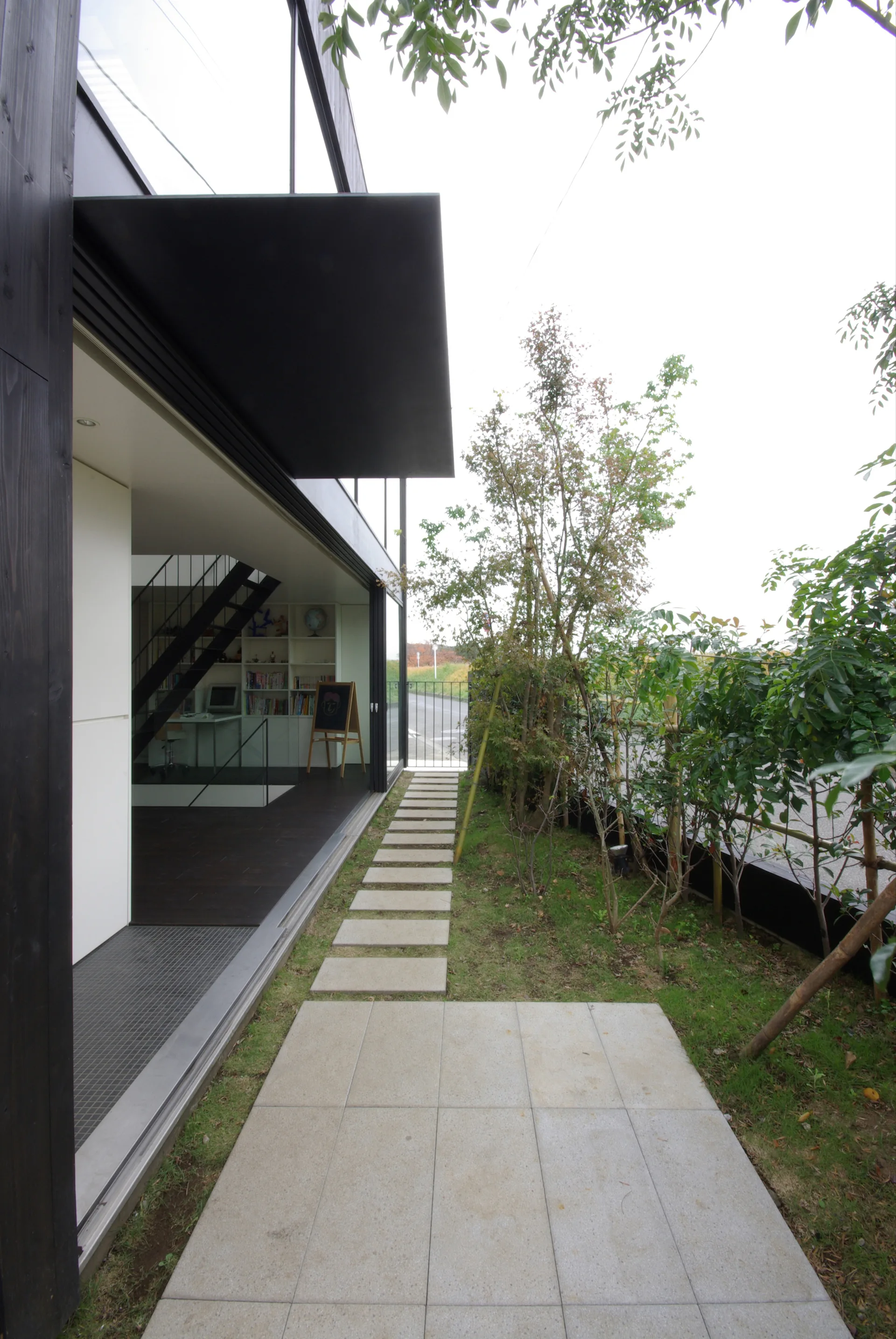 A modern black house features an open sliding glass wall showing a study with a black staircase next to a concrete garden stepping stone path.
