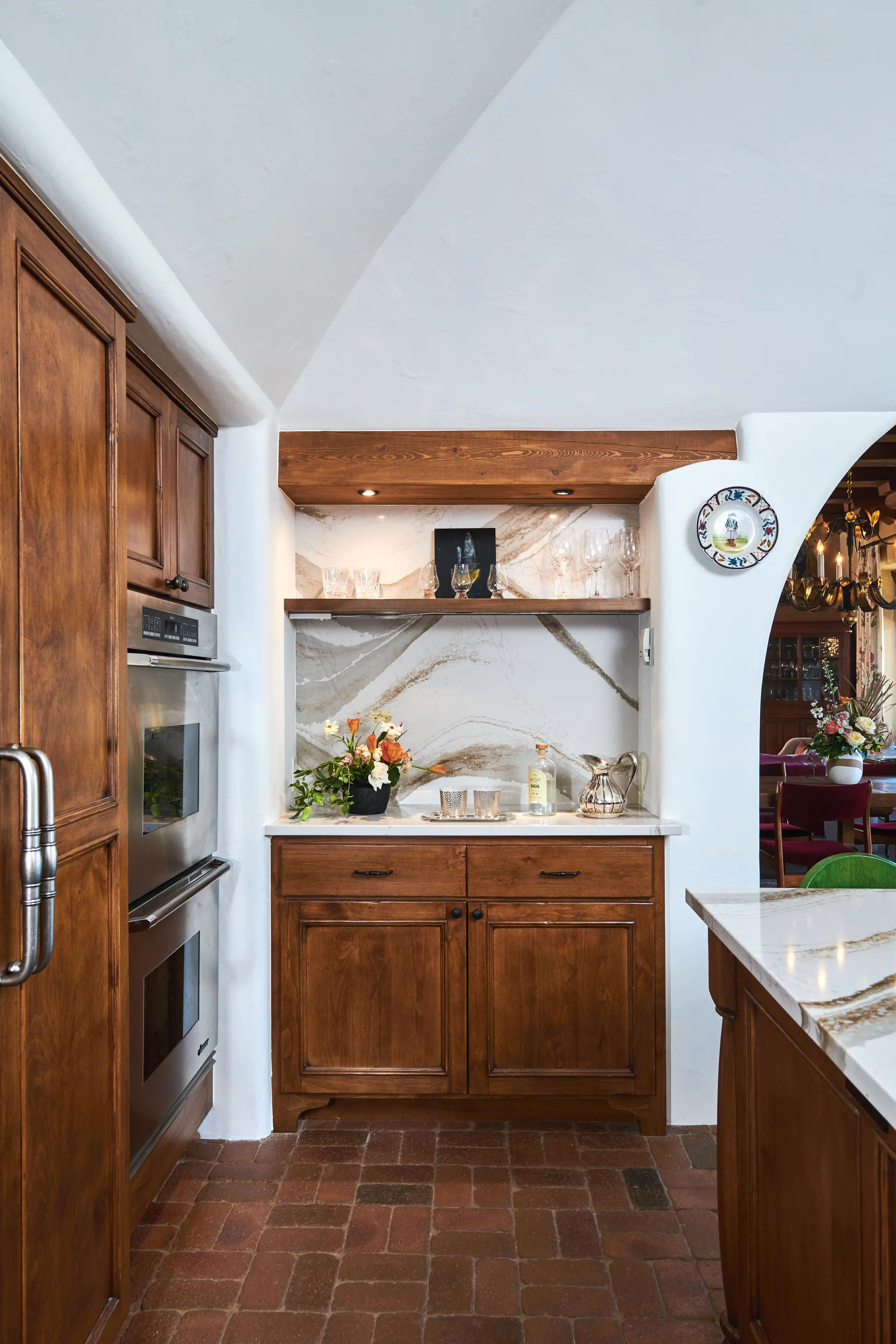 A rustic built-in wood beverage bar featuring a heavily veined stone backsplash and countertop, wooden cabinets, and terracotta brick tile flooring.