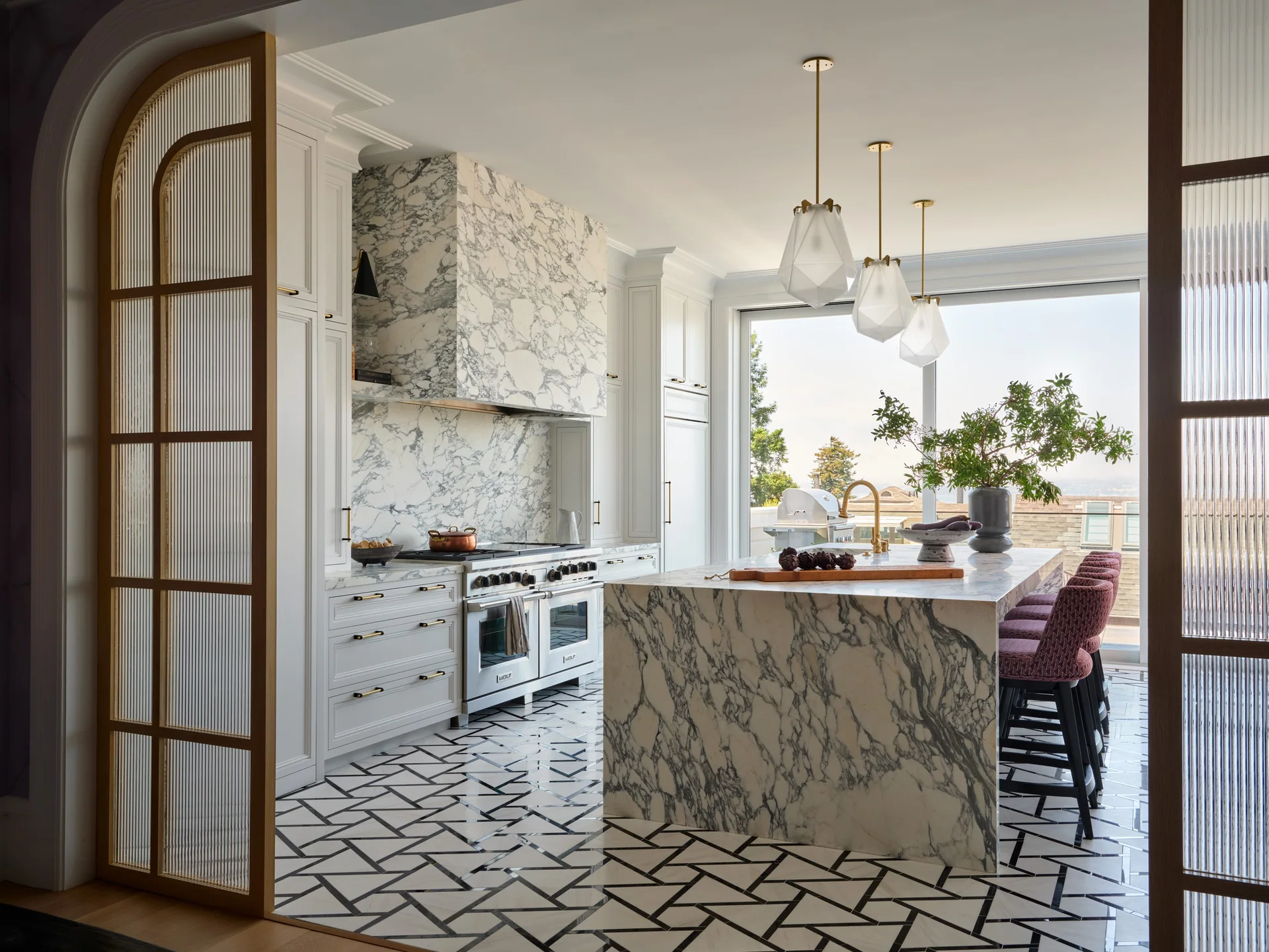 A modern kitchen featuring a heavily veined marble waterfall island, matching range hood, geometric floor tile, and a wooden archway with fluted glass.