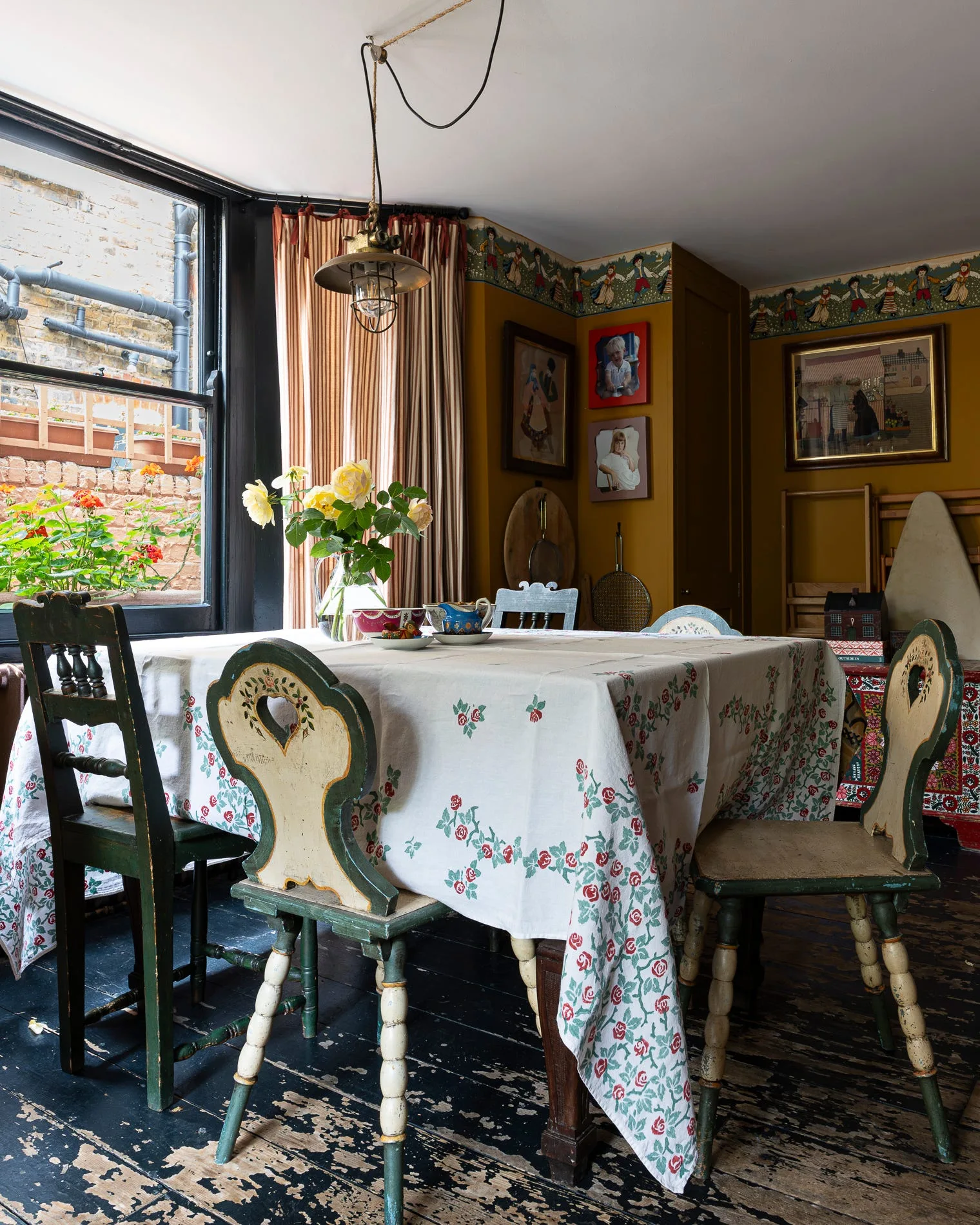 A rustic dining table covered in a floral tablecloth is surrounded by painted wooden chairs in a room with mustard walls and a decorative border.