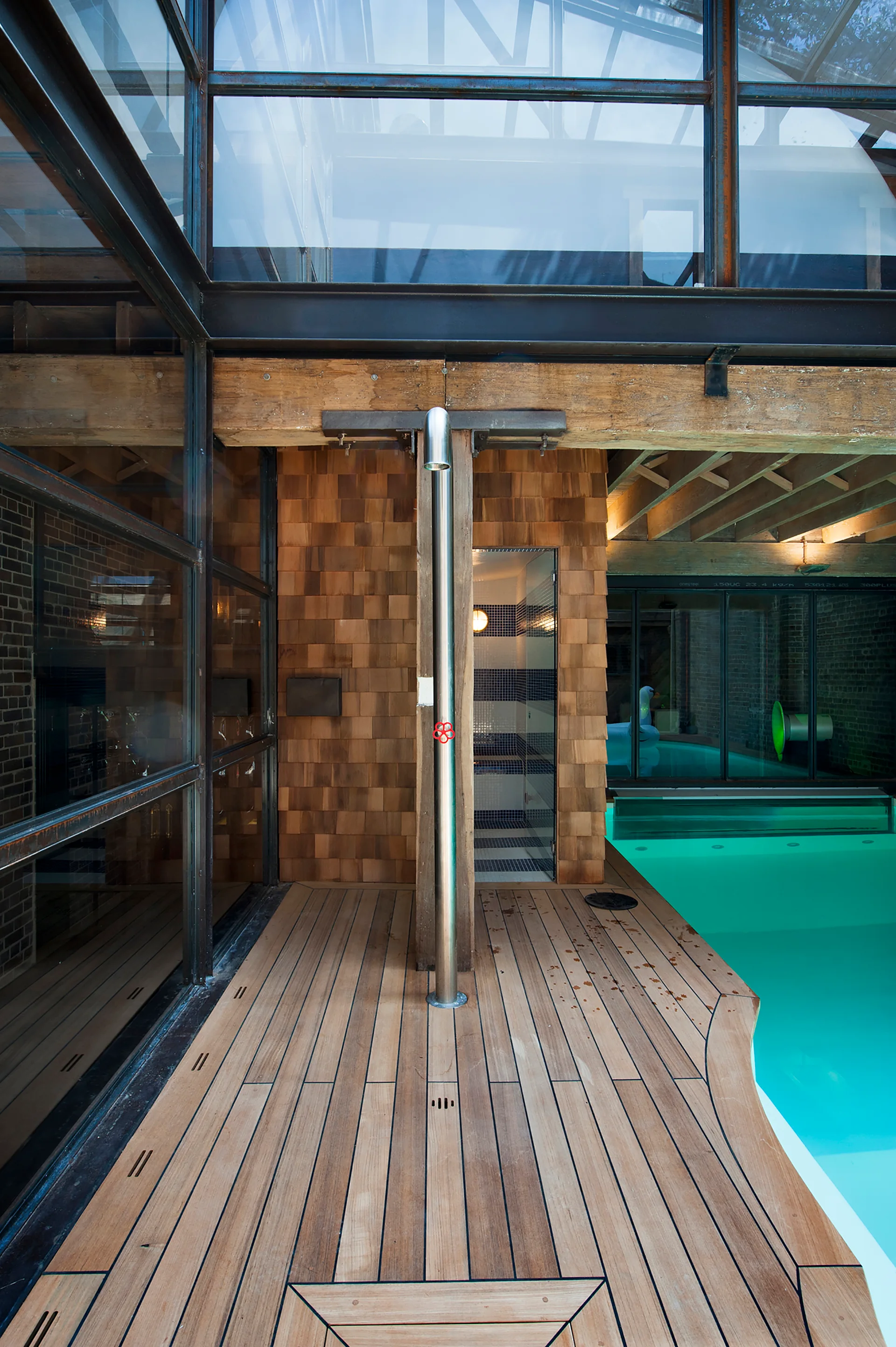 An indoor-outdoor pool deck features a freestanding stainless steel shower, natural wood decking, cedar shingle siding, and a turquoise pool.