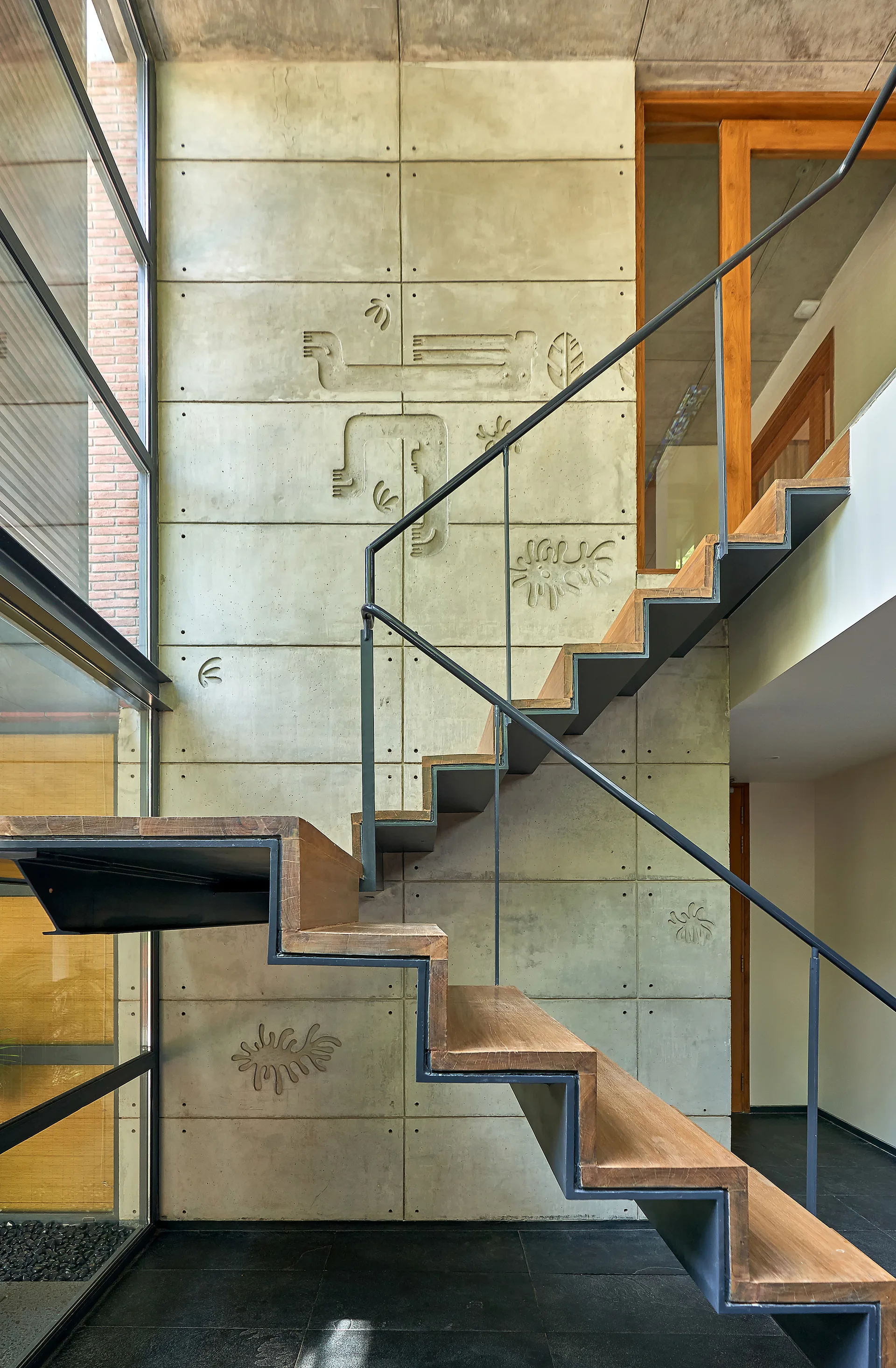 A modern floating staircase with wood treads and a black steel frame stands in front of a concrete block wall carved with abstract drawings.