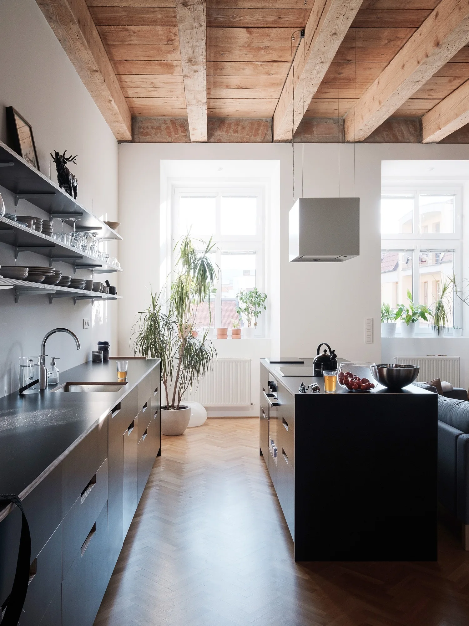 A modern kitchen with matte black base cabinets, a central island, exposed rustic ceiling beams, and light herringbone wood floors.