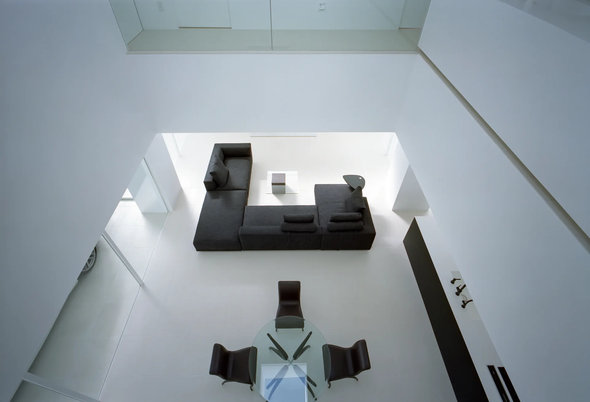 A high-angle view looking down into a stark white living area with a large charcoal gray sectional sofa and a glass-topped dining table.