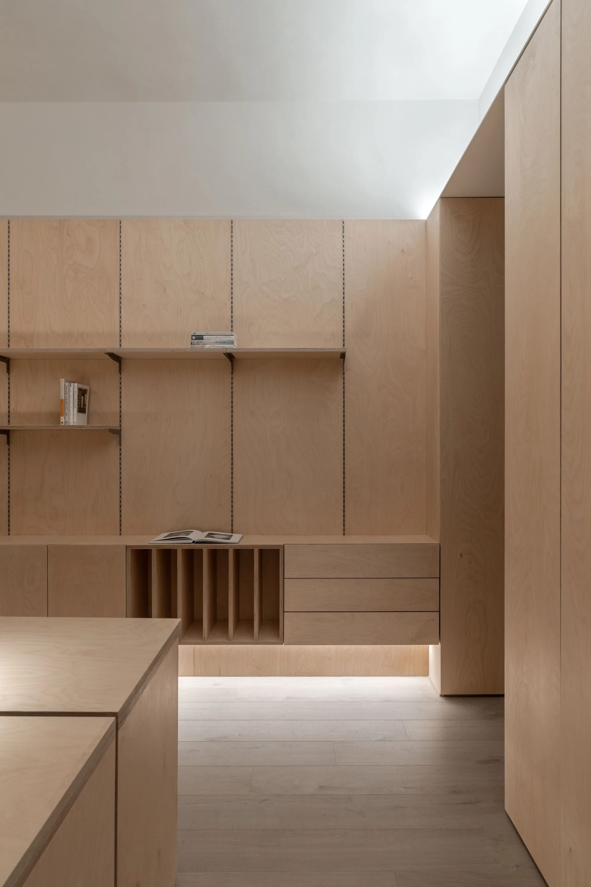 A minimalist room features custom plywood wall paneling, built-in lower cabinets with a slotted organizer, adjustable wall shelves, and floor lighting.