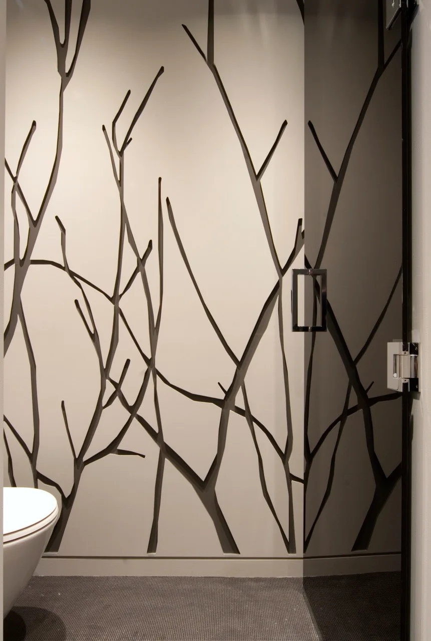 A modern powder room featuring a wall with a cutout branch motif, a dark glass door with a geometric metal handle, and a grey tiled floor.