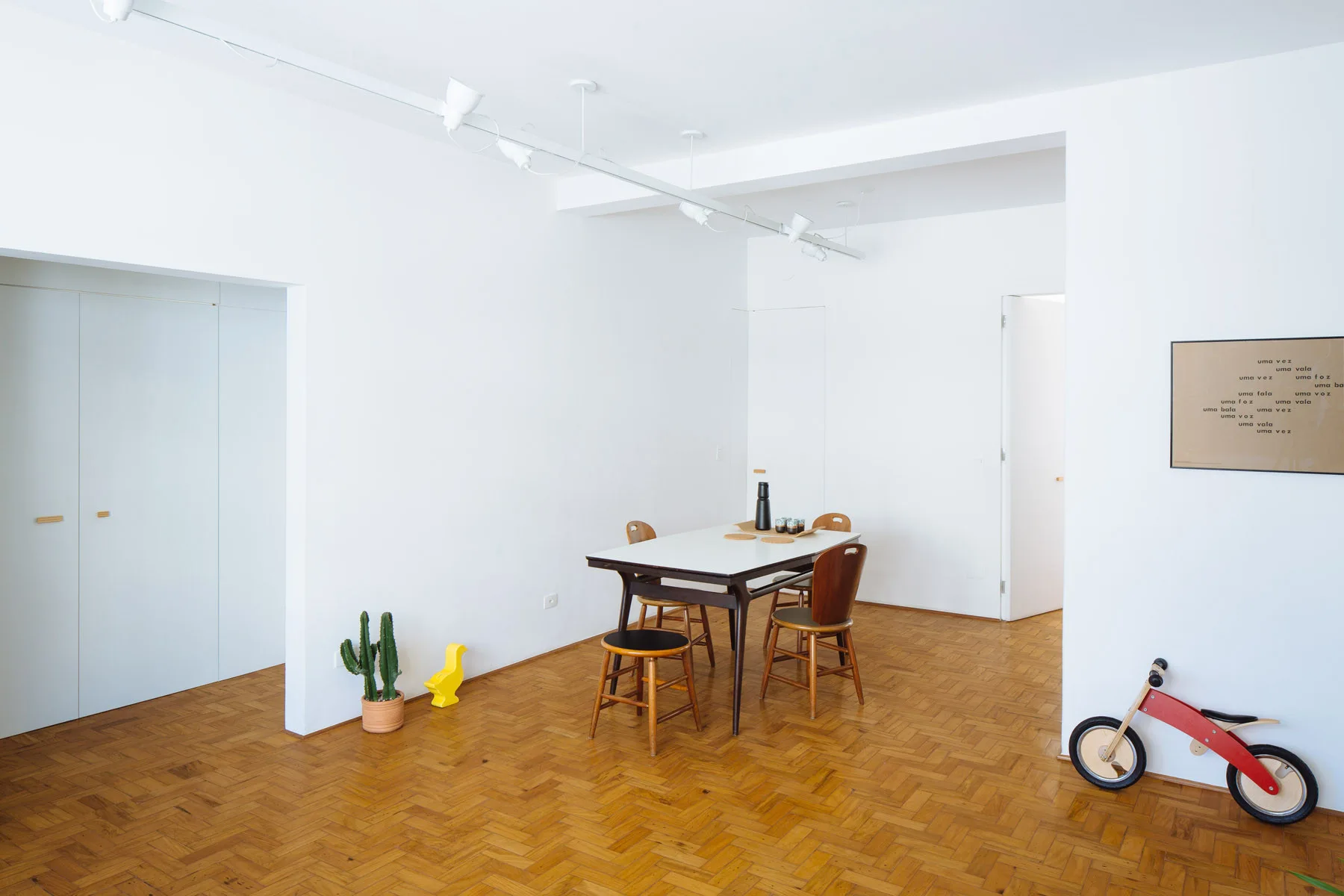 A minimalist dining room features a white-topped wooden table and mismatched chairs on herringbone parquet flooring against plain white walls.