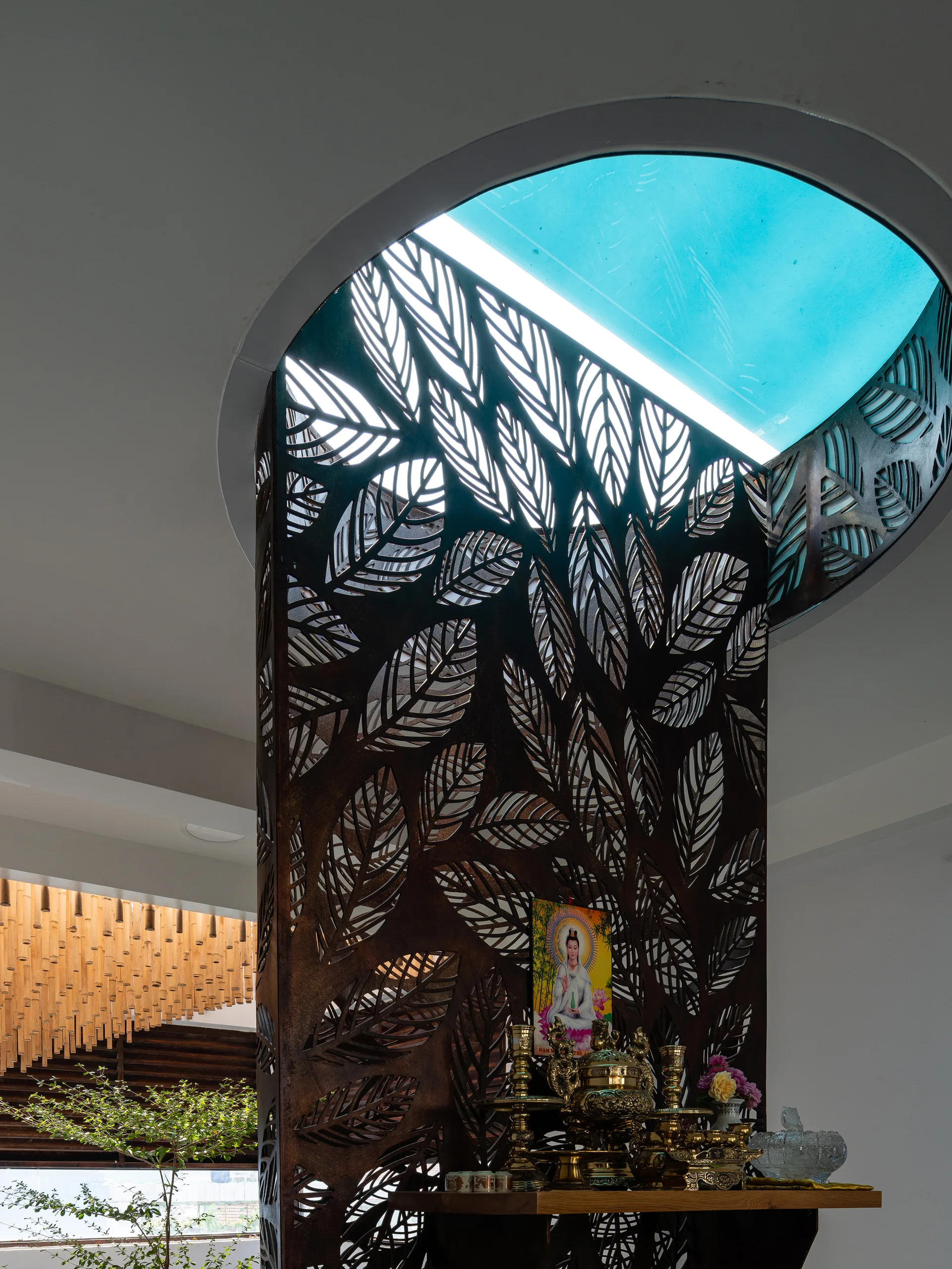 A dark metal laser-cut leaf pattern screen stands beneath a circular skylight, serving as a backdrop for a wooden altar with brass items.