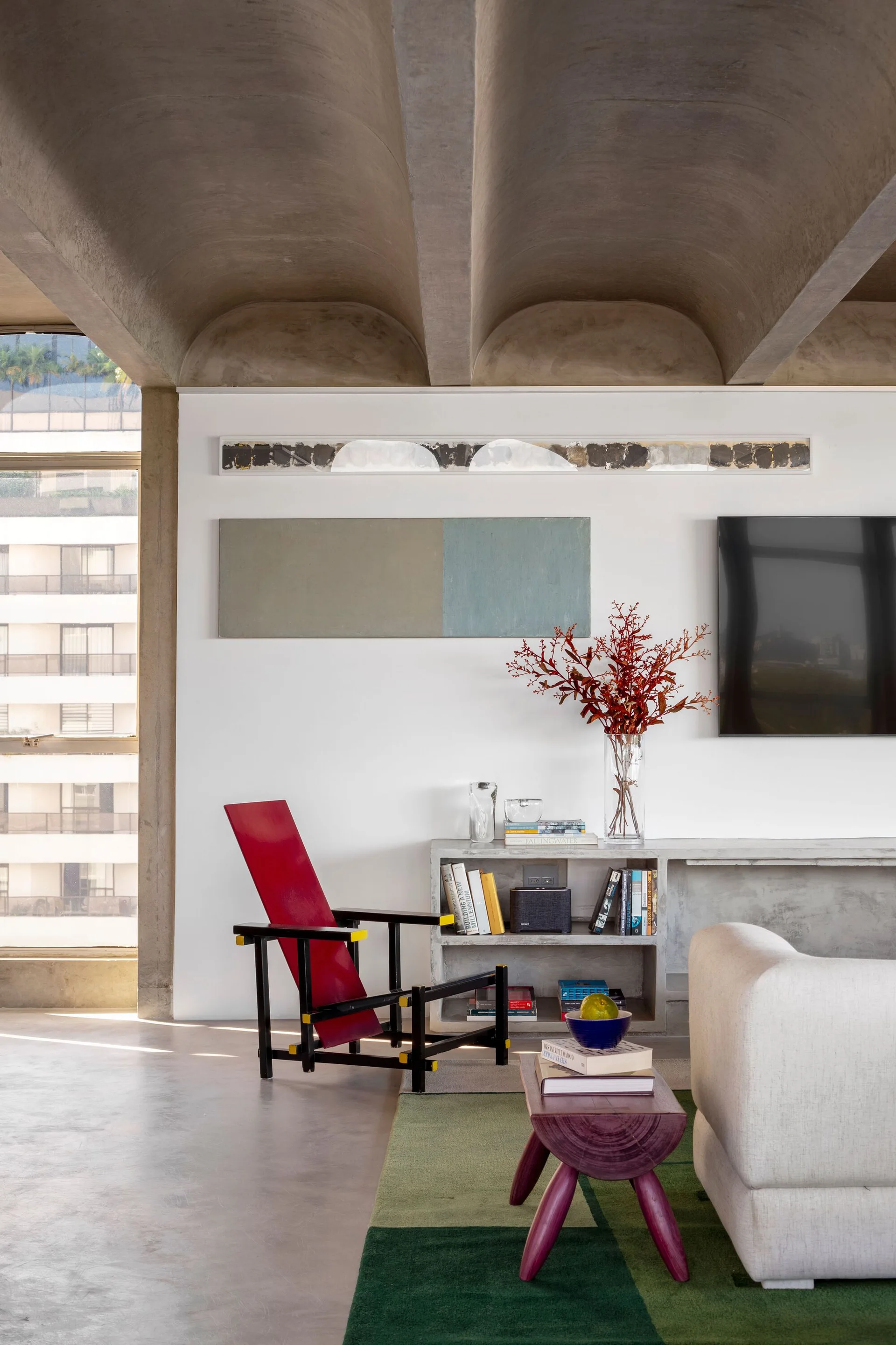 A modern living space features a gray concrete vaulted ceiling, a red accent chair, and a low concrete storage console beside a green rug.