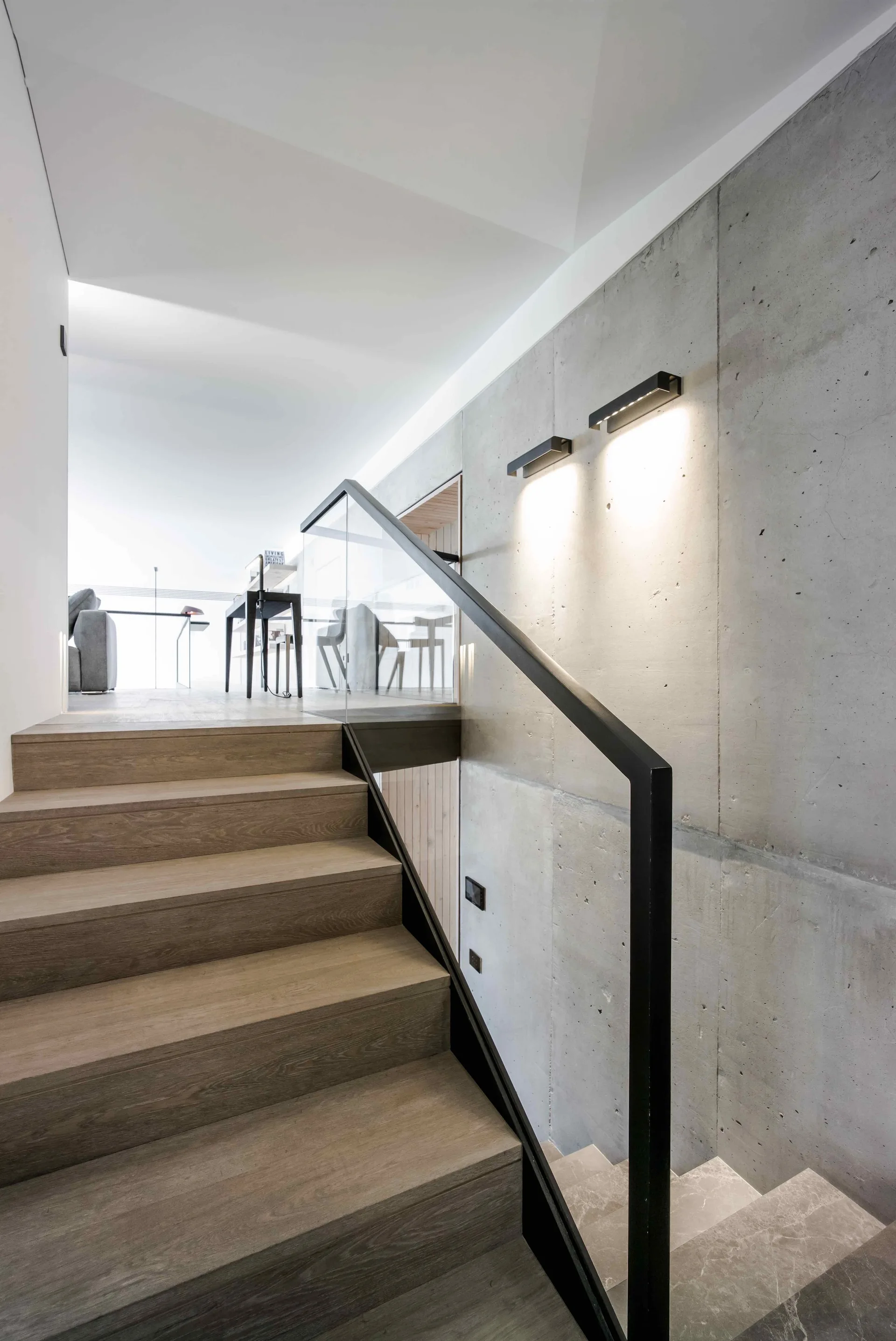 A modern staircase features warm wood treads, a glass railing with a black handrail, and a tall exposed concrete wall illuminated by sleek black wall lights.