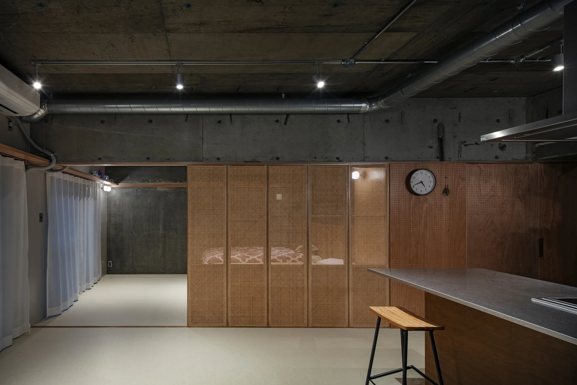 An industrial interior features raw concrete ceilings with ductwork, woven cane partition panels, a pegboard wall, and a stainless steel kitchen island.