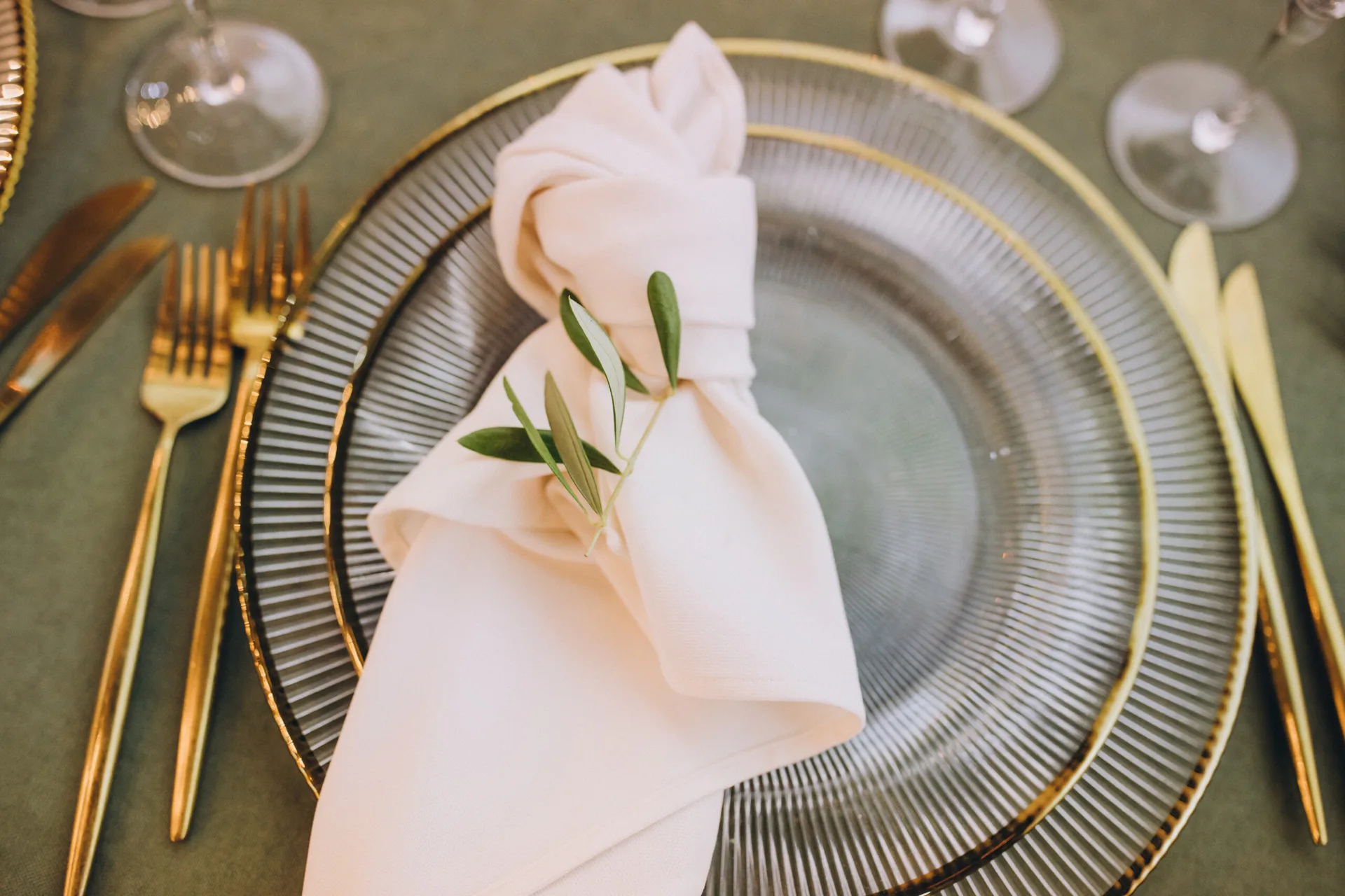 A tabletop place setting with a ribbed glass plate, a gold rim, a knotted cream cloth napkin with an olive sprig, and gold cutlery.