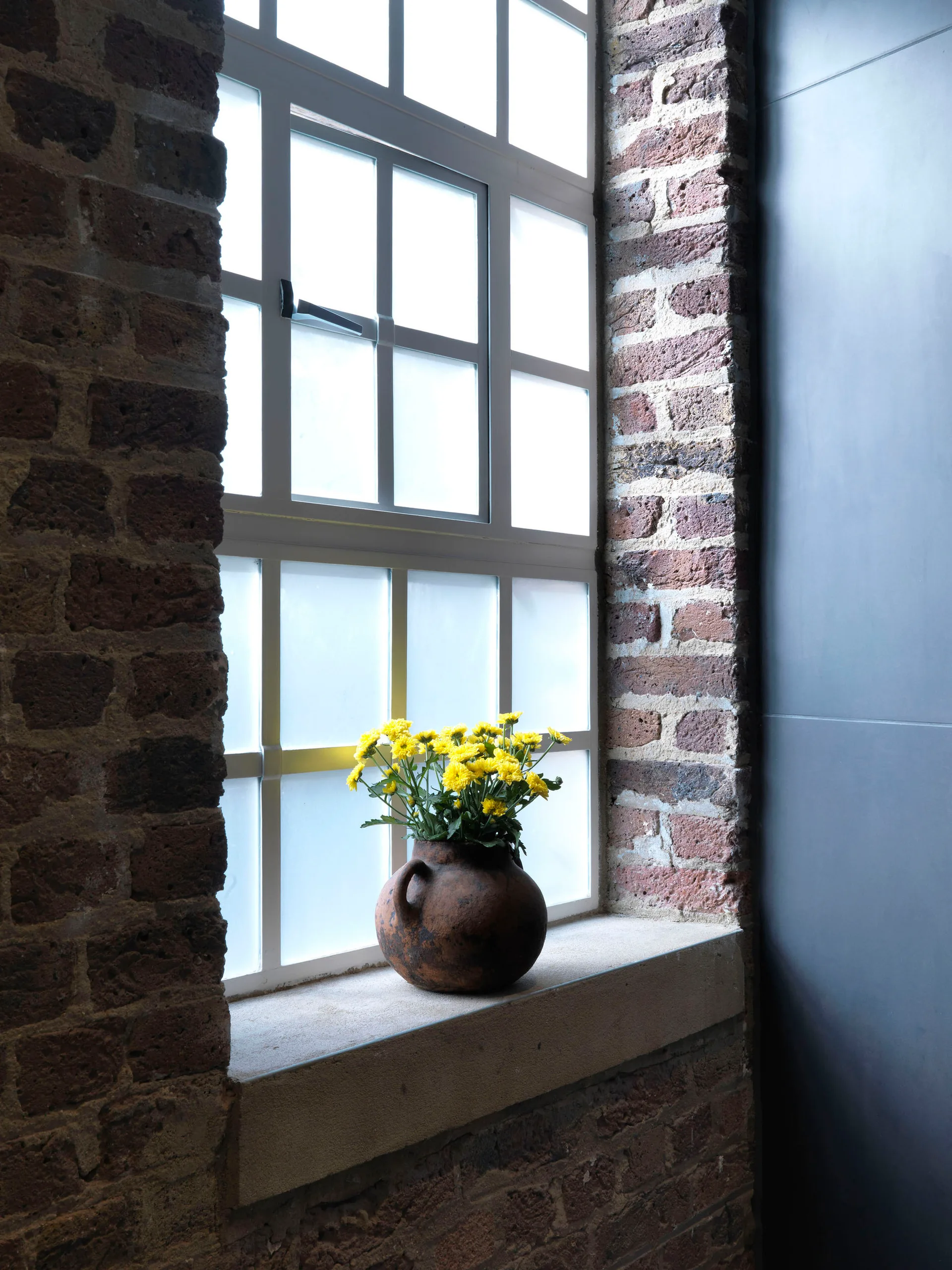 A rustic brown clay vase filled with bright yellow flowers sits on a stone sill of a grid-paned window surrounded by exposed red brick walls.