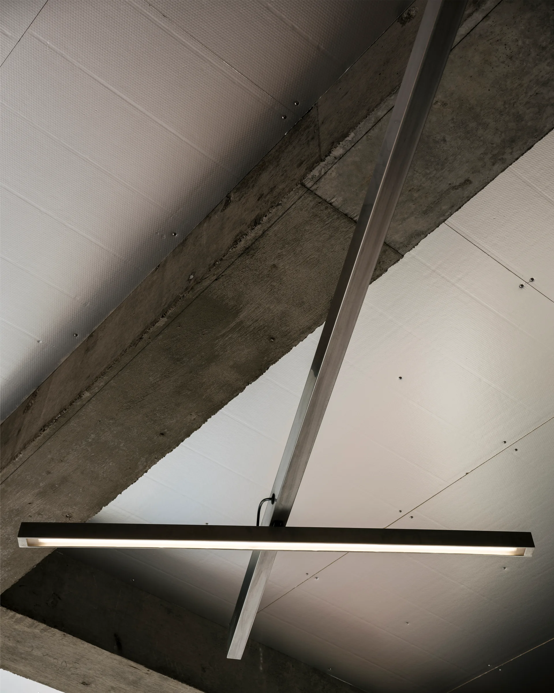 A low-angle shot of a minimalist linear pendant light hanging beneath an exposed concrete structural beam and white ceiling panels.