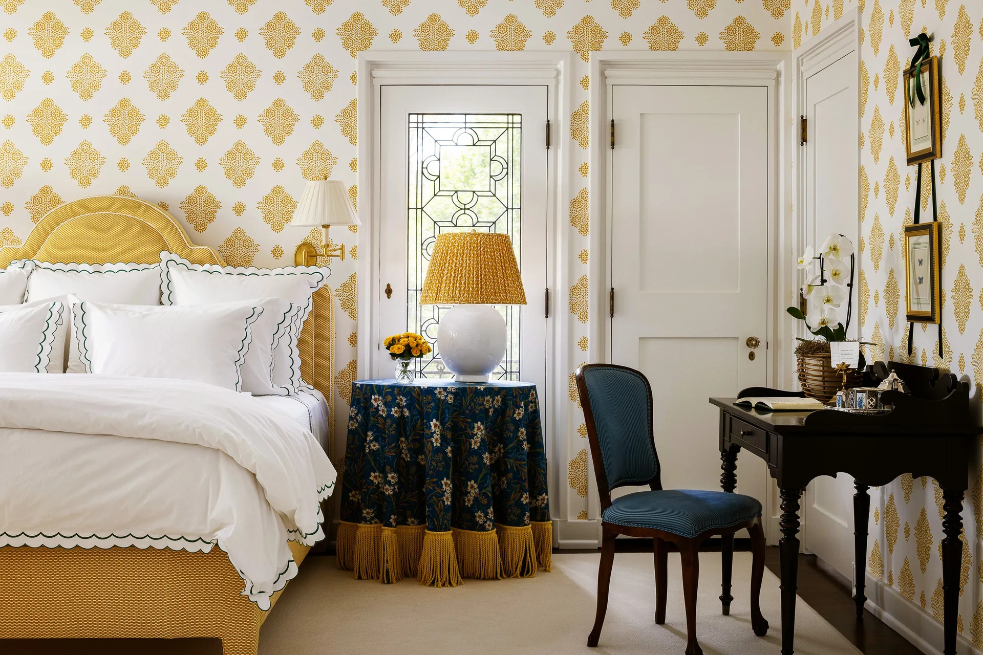 A traditional bedroom features yellow patterned wallpaper, an upholstered yellow bed, a fringed skirted side table, and a dark wood writing desk.