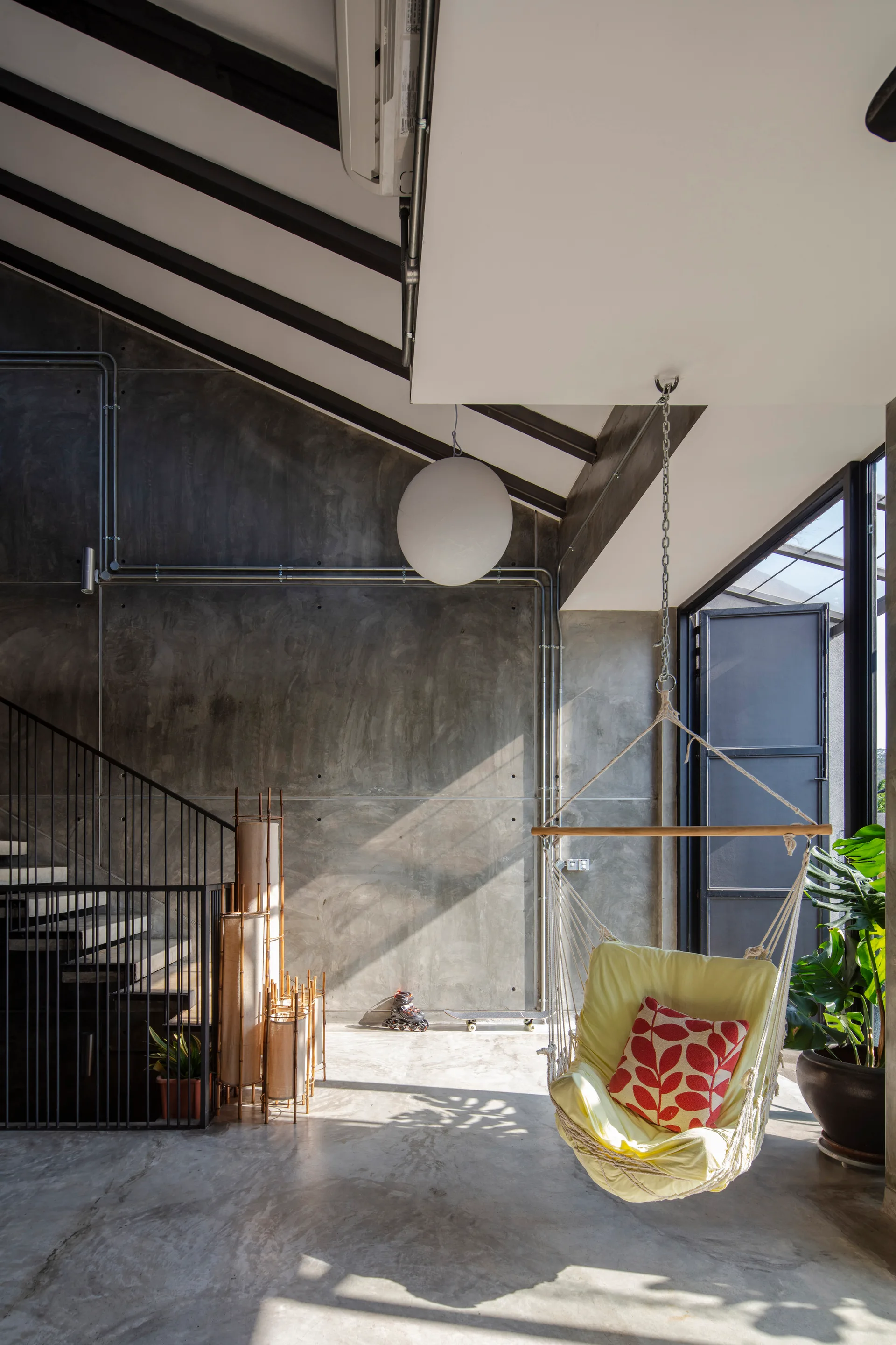 An industrial living area features raw concrete walls, a black metal staircase, and a yellow hanging swing chair suspended from the ceiling.