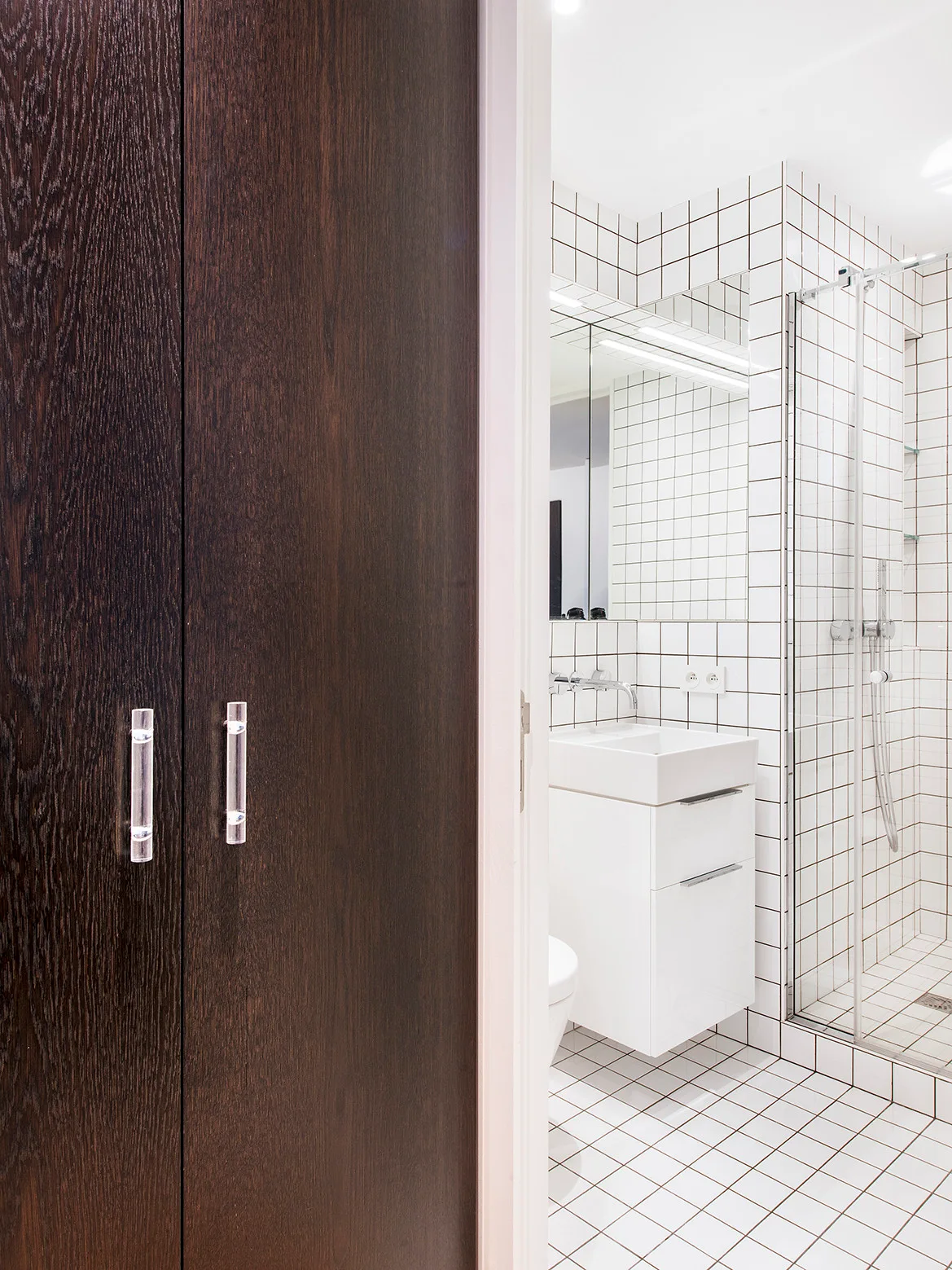 A view through a dark wood doorway into a modern bathroom featuring white grid-patterned tiles, a wall-mounted vanity, and a glass-enclosed shower.