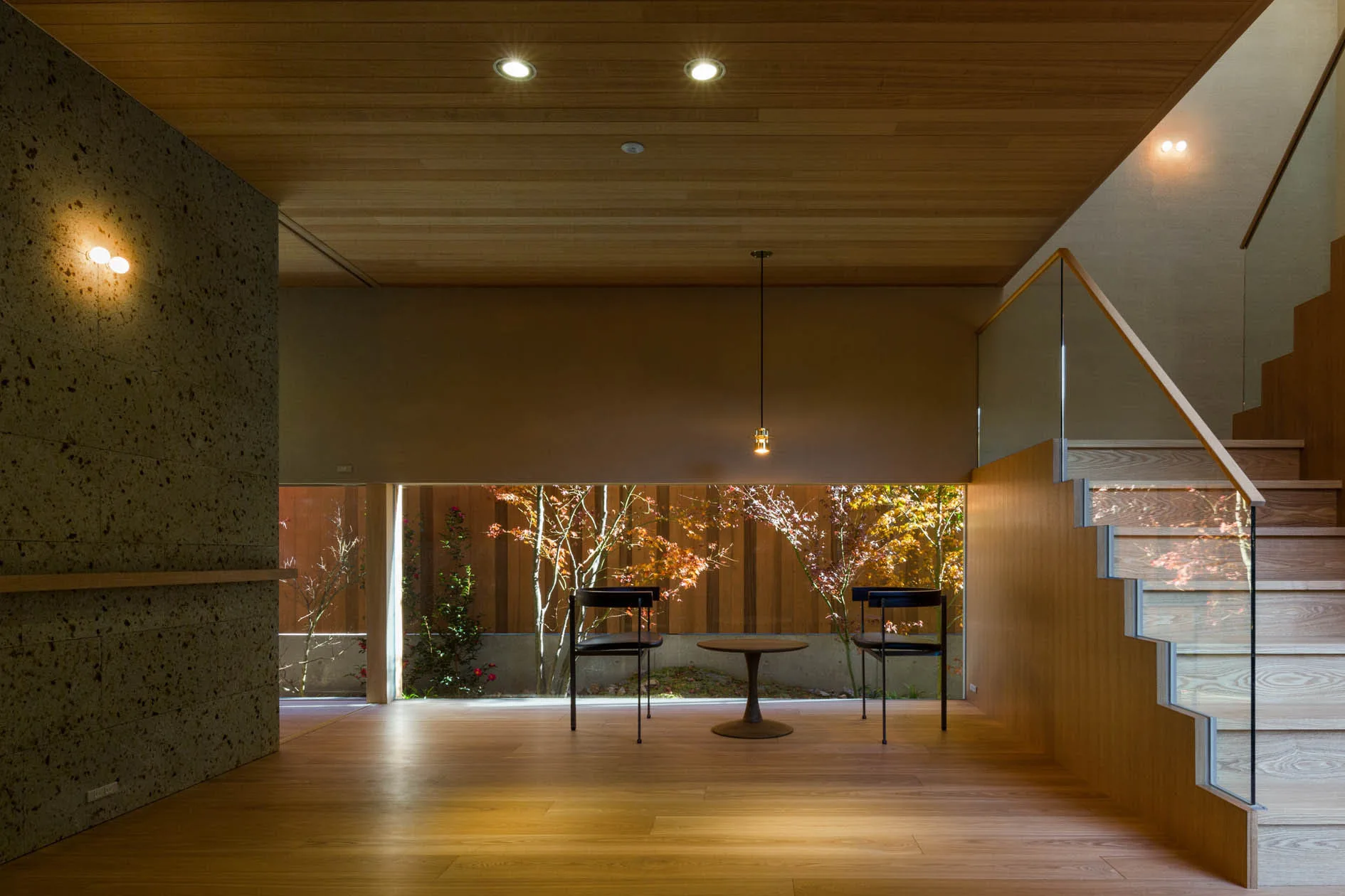 A minimalist wooden room features a low horizontal window framing garden trees, a staircase with a glass railing, and two dark chairs.
