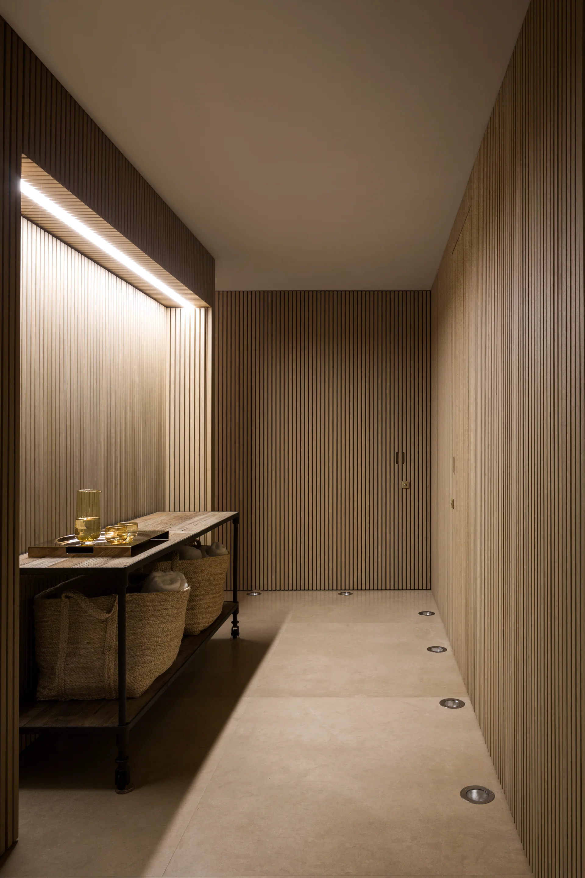 A modern hallway features vertical slatted wood walls, a built-in illuminated alcove with a console table, and recessed floor spotlights.
