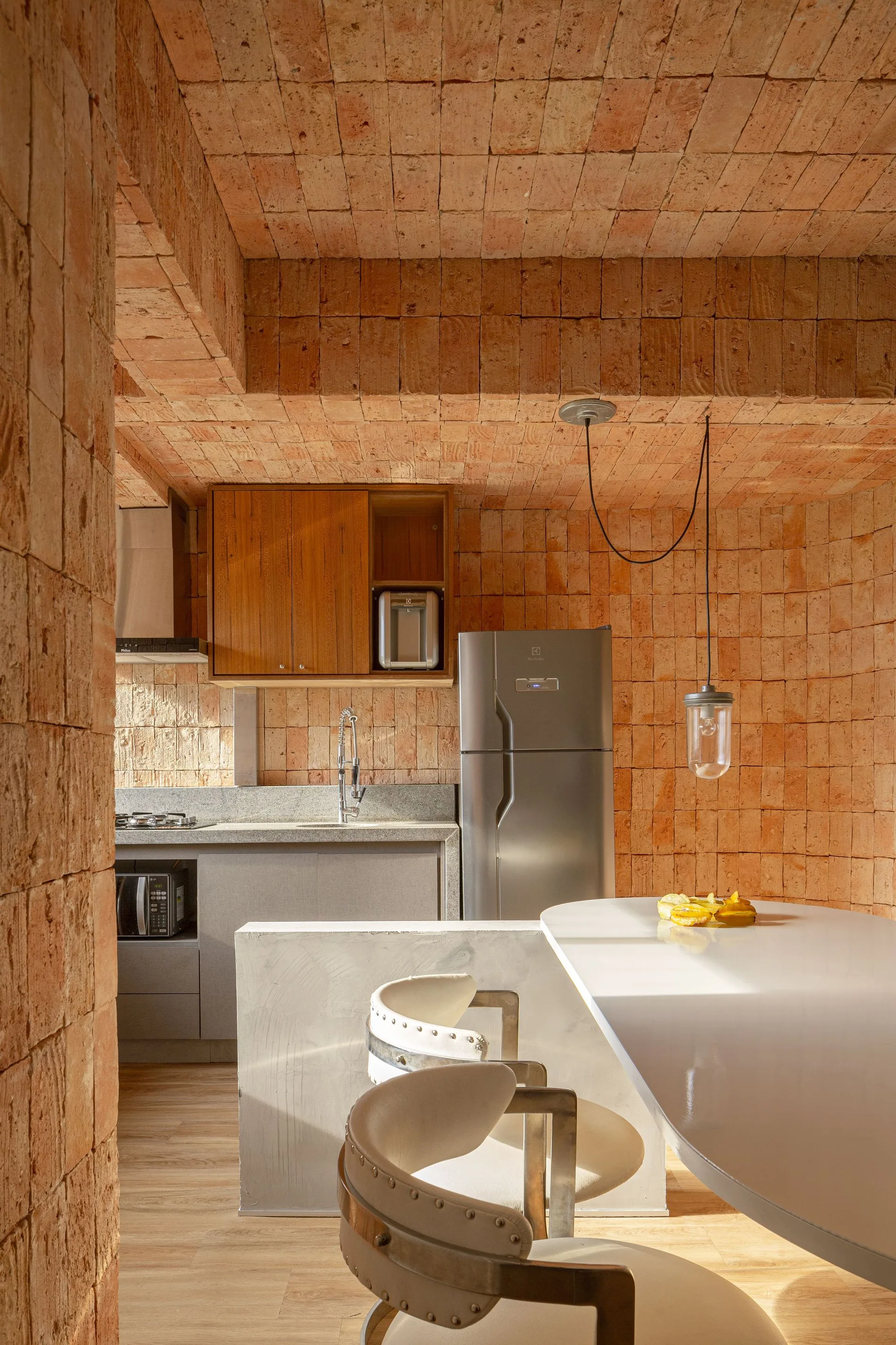 A compact kitchen features terracotta brick walls and ceiling, a white breakfast bar with curved stools, and stainless steel appliances.