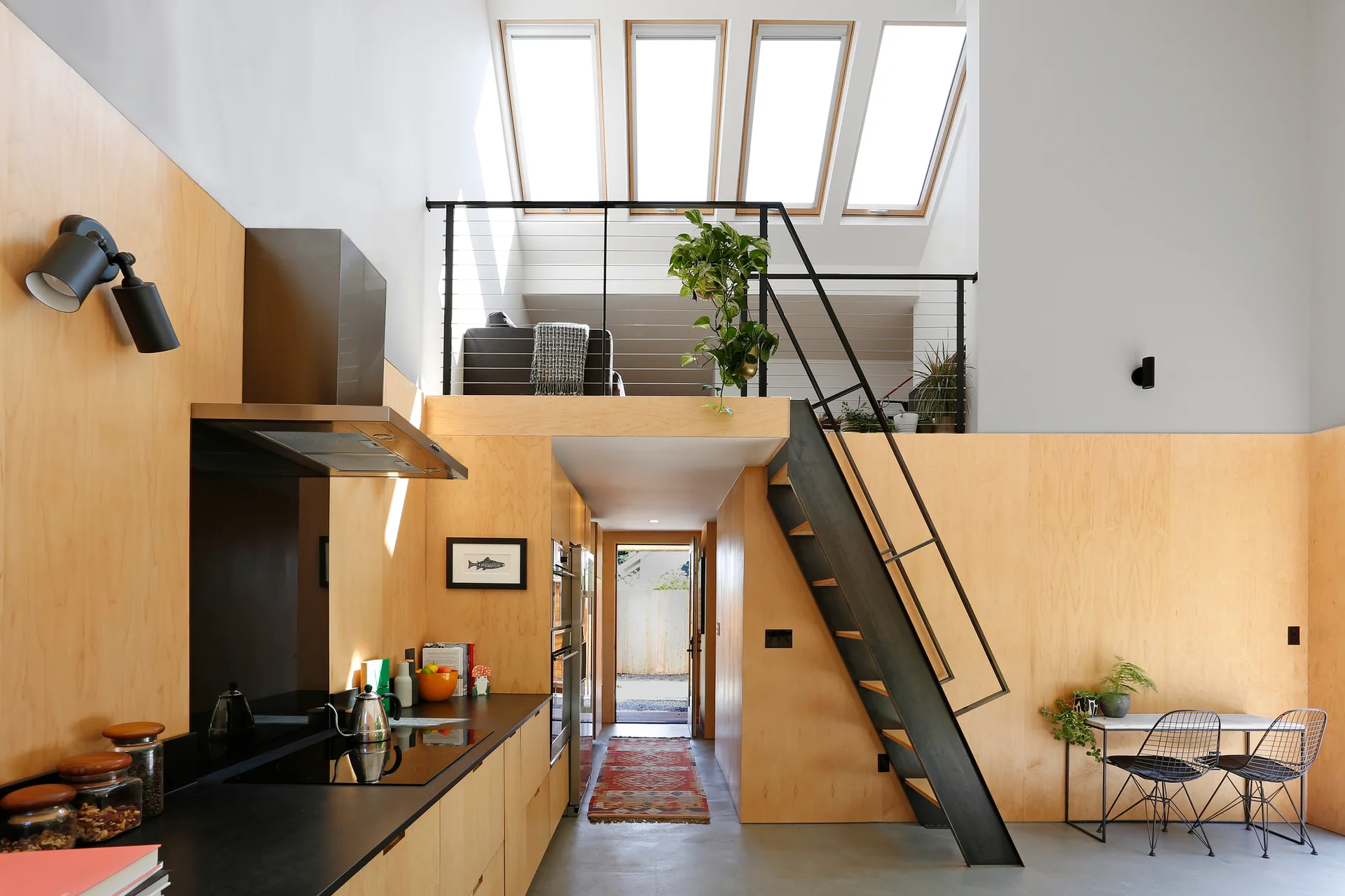 A minimalist kitchen features light plywood-clad walls, a black metal staircase leading to a mezzanine, and a row of four slanted ceiling skylights.