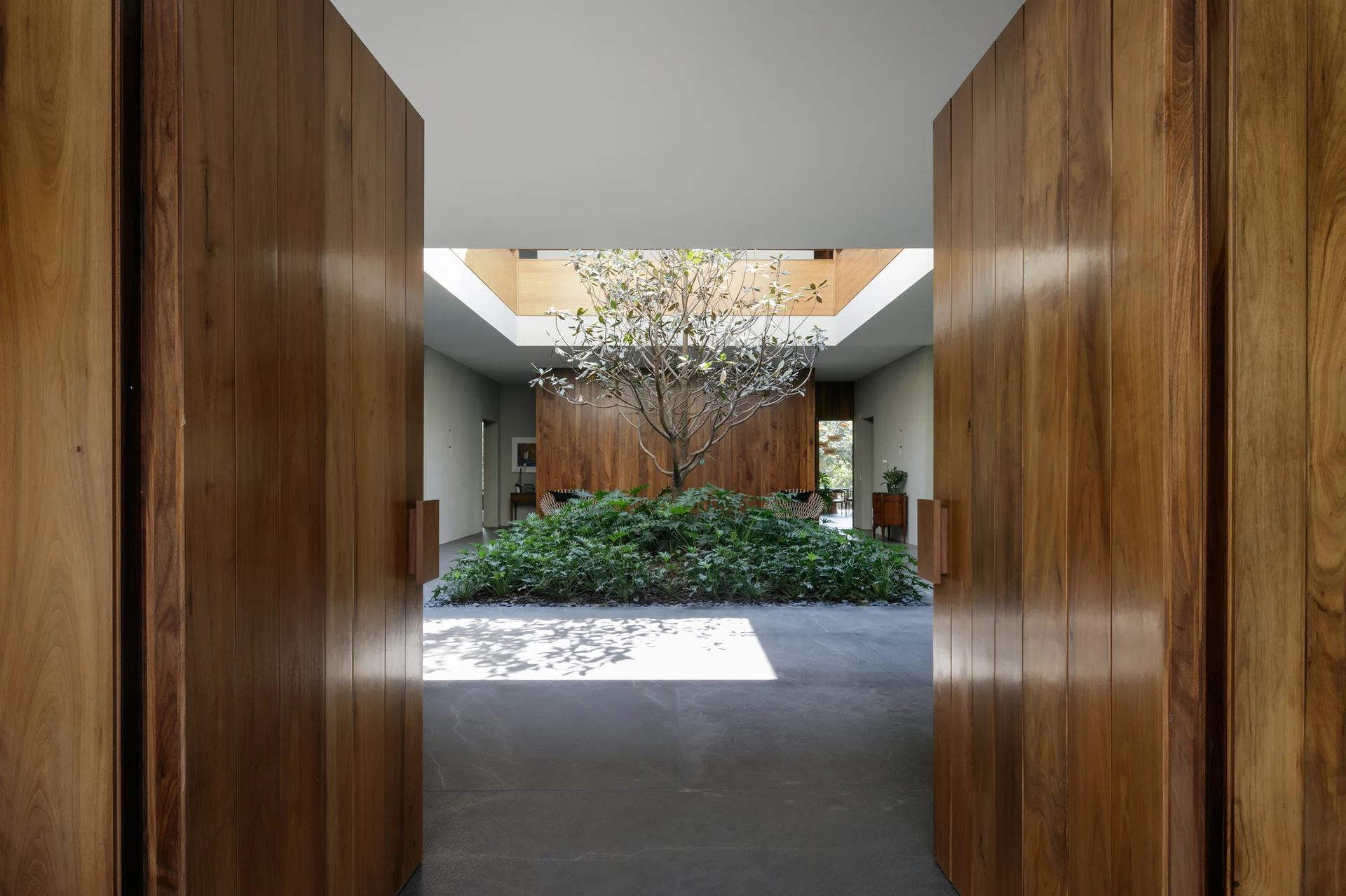 Large open wooden entry doors frame a modern indoor courtyard with a central tree, green foliage, a skylight, and grey stone flooring.