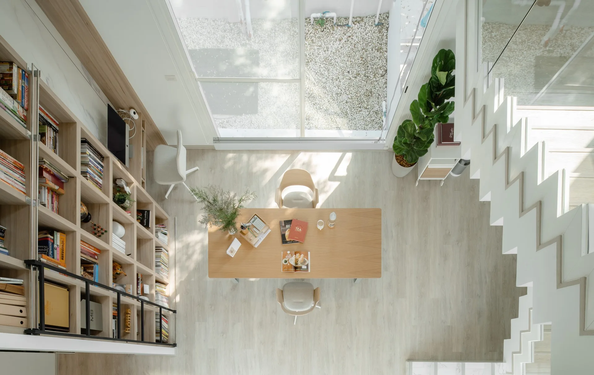 Overhead view of a minimalist dining room with a light wood table, a tall wooden bookcase with a ladder, and a white geometric staircase.
