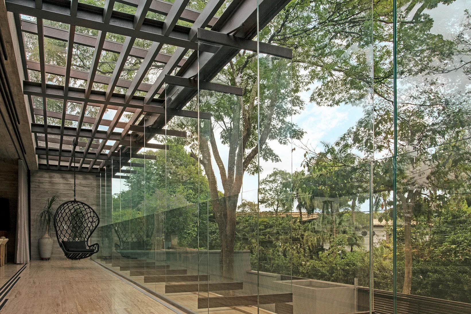 A modern outdoor terrace features a dark metal pergola ceiling, polished stone floors, a black hanging chair, and frameless glass walls looking out at trees.