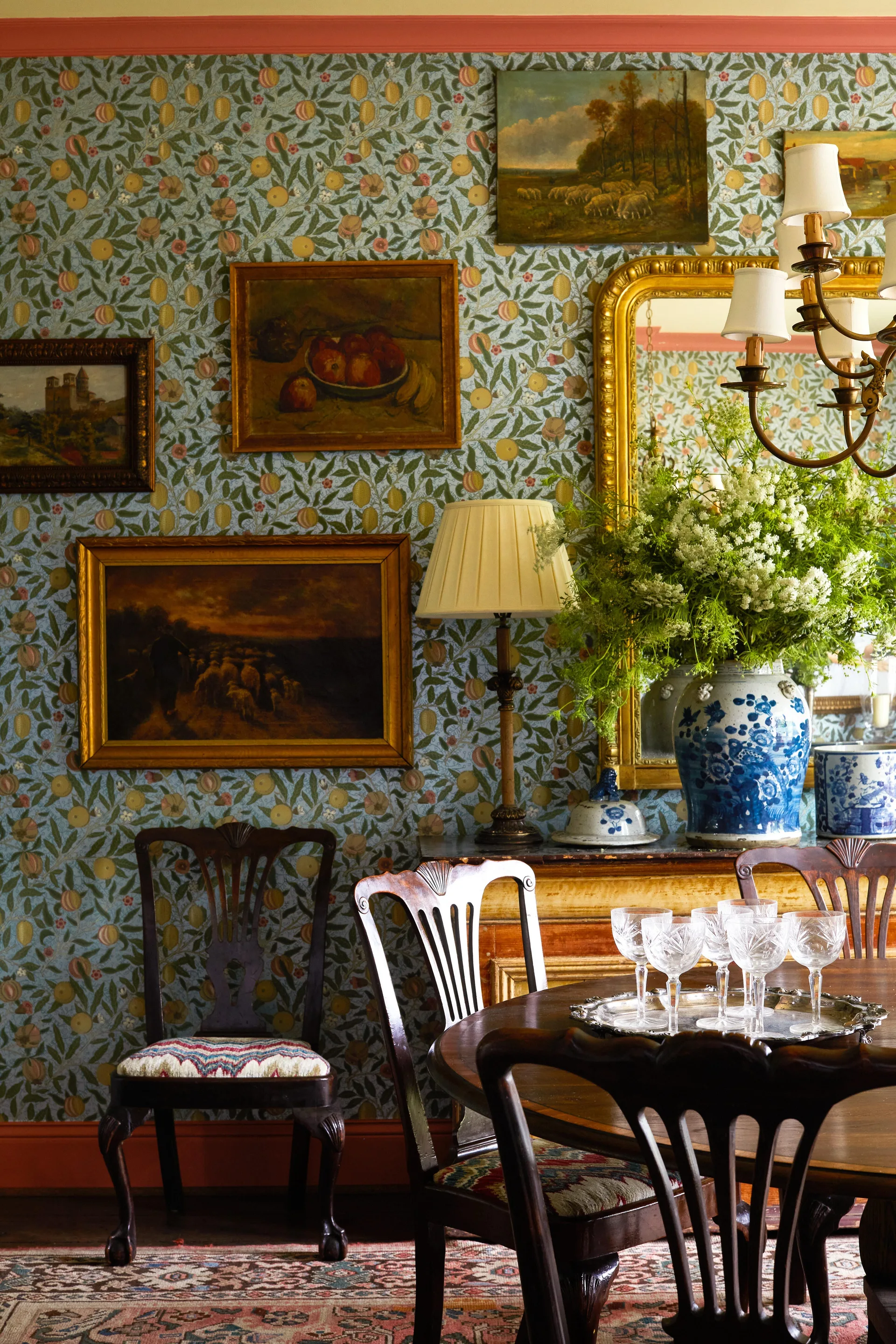 A traditional dining room features blue-green floral wallpaper adorned with gold-framed paintings, a wooden table with crystal glasses, and a gilt mirror.