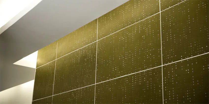 A wall clad in olive green perforated metal panels in a grid pattern, highlighted by recessed ceiling lights.