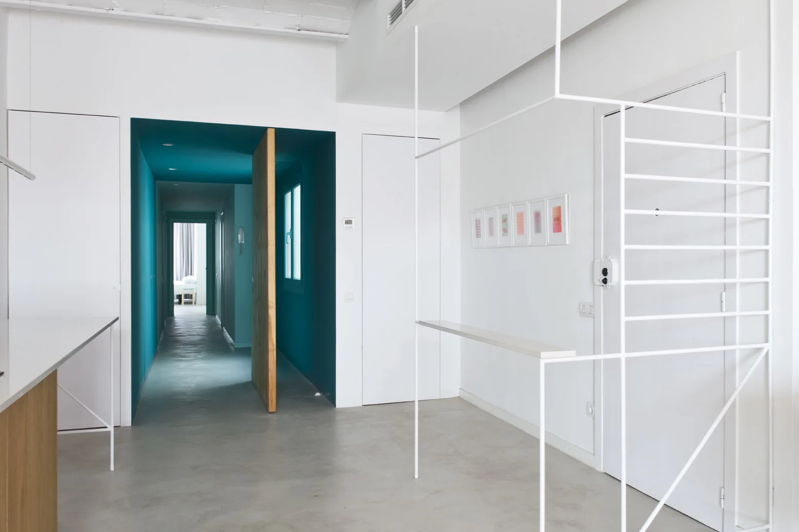 A minimalist white entryway featuring a slender white metal partition, smooth concrete flooring, and an open doorway leading into a vibrant teal hallway.