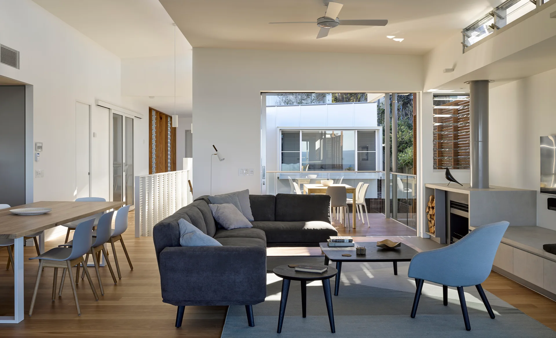 An open-plan living room features a dark gray sectional sofa, a light blue armchair, a wooden dining table, and sliding glass doors opening to an outdoor deck.