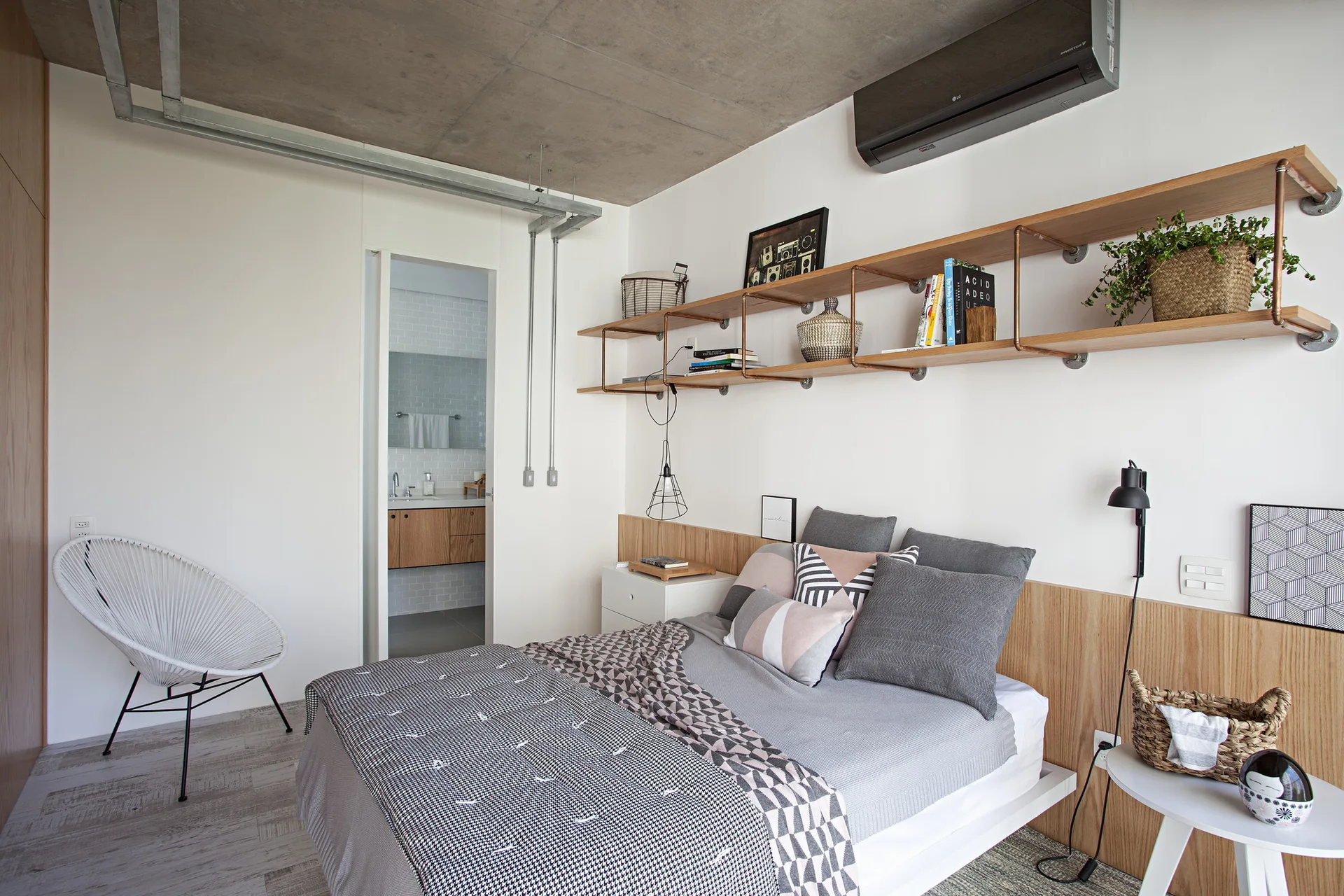 A modern industrial bedroom features a bed with geometric bedding, a wood headboard panel, and open copper-pipe wall shelving beneath a concrete ceiling.