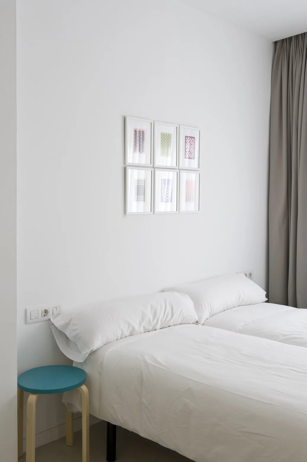 A minimalist bedroom with white bedding, a small wood and blue bedside stool, and a grid of six small framed prints on the white wall.