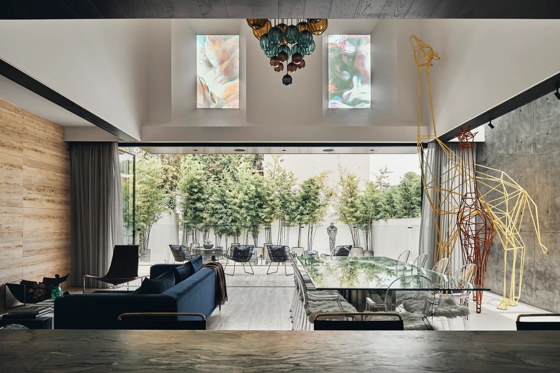 A modern open-plan dining and living space features a glass-top table with clear acrylic chairs, a blue sofa, and tall wire giraffe sculptures.