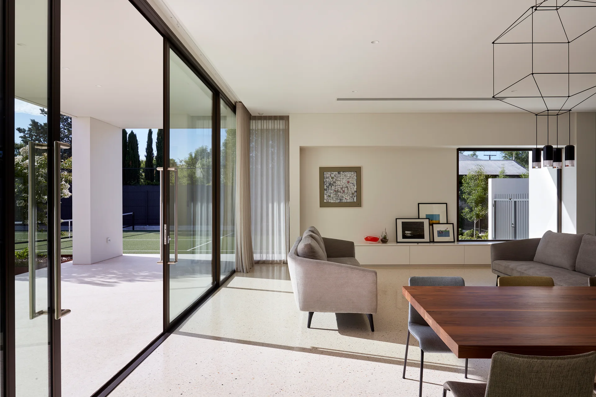 A modern open-plan living room features speckled terrazzo flooring, two grey sofas, a wooden dining table, and large sliding glass doors opening to a patio.