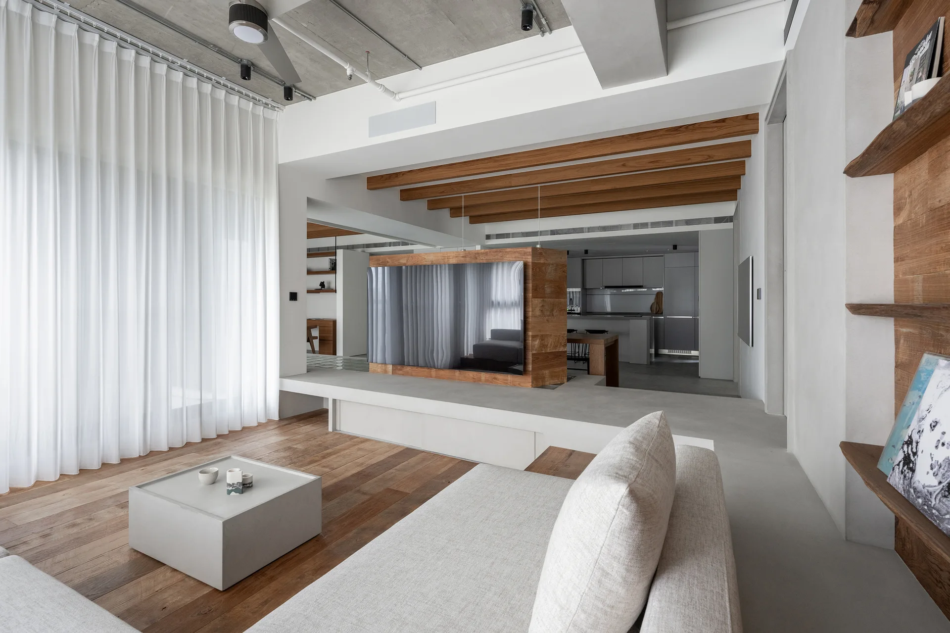 A minimalist living room featuring a concrete ceiling, a central wooden TV partition, a low grey sofa, and sheer white curtains.