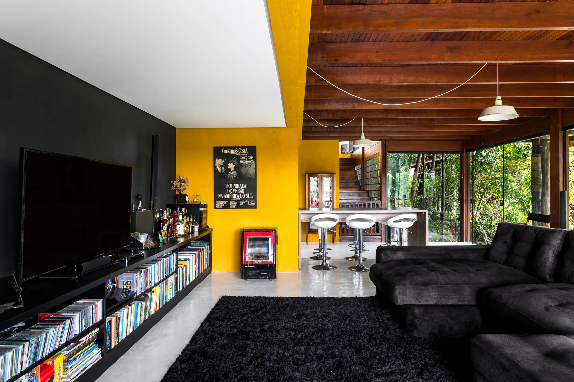A modern living room features a yellow accent wall, a black media unit under a television, a dark sectional sofa, and an exposed wood beam ceiling.