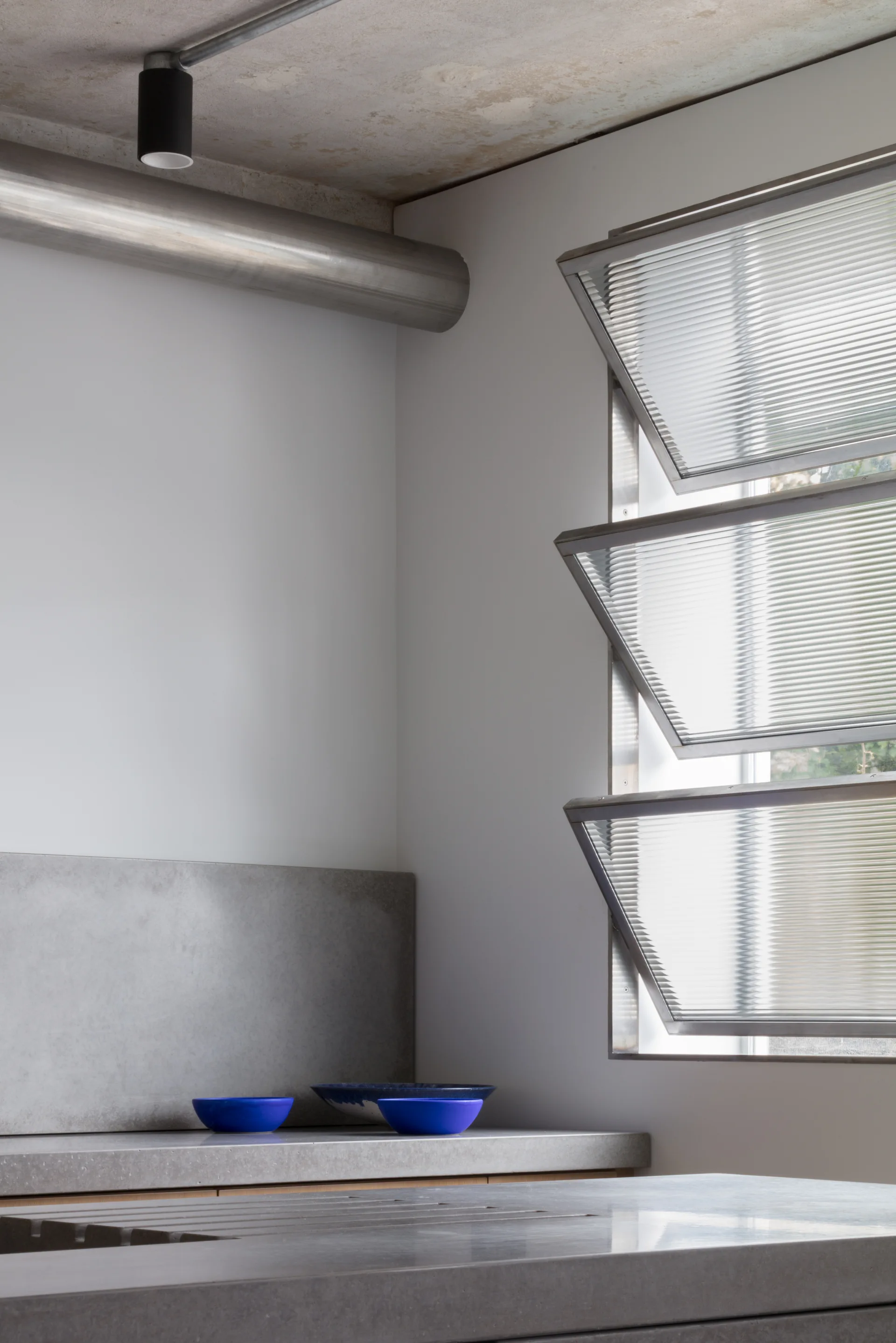 An industrial kitchen corner featuring concrete countertops, a smooth concrete backsplash, small cobalt blue bowls, and louvered glass windows.