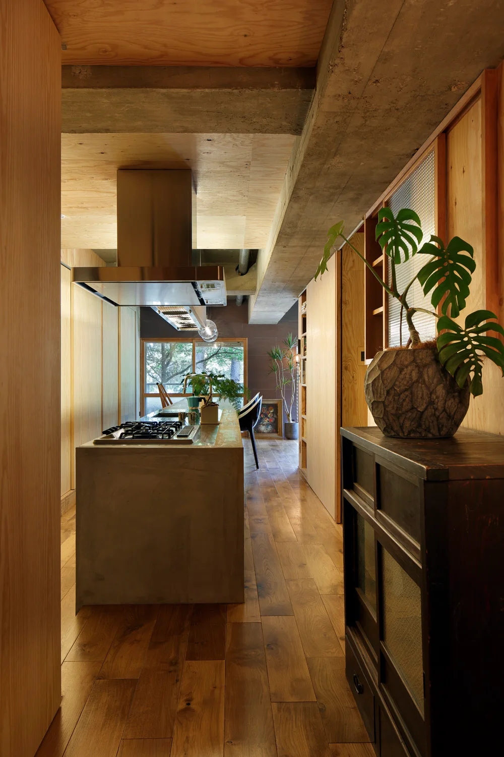 A modern industrial kitchen with a central concrete island, stainless steel range hood, warm plywood walls, and exposed concrete ceiling beams.